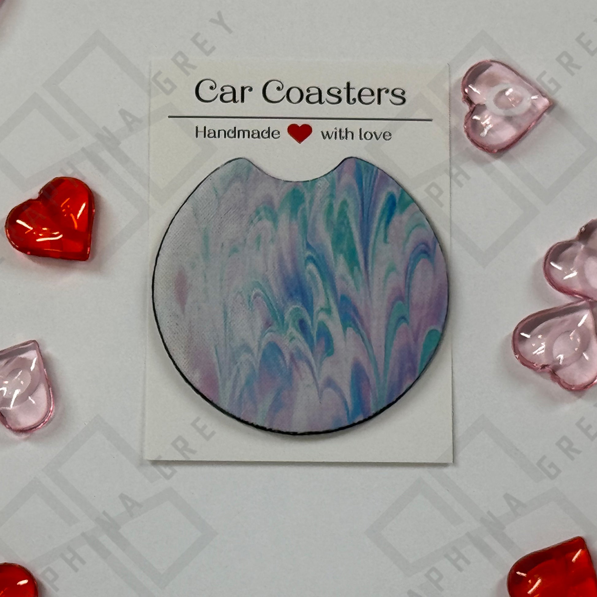 A single car coaster featuring a blue tie-dye design with swirling patterns in shades of blue, purple, and white. The coaster is displayed with a label that reads ‘Car Coasters - Handmade with love.’ The background is adorned with red and pink heart-shaped decorations.