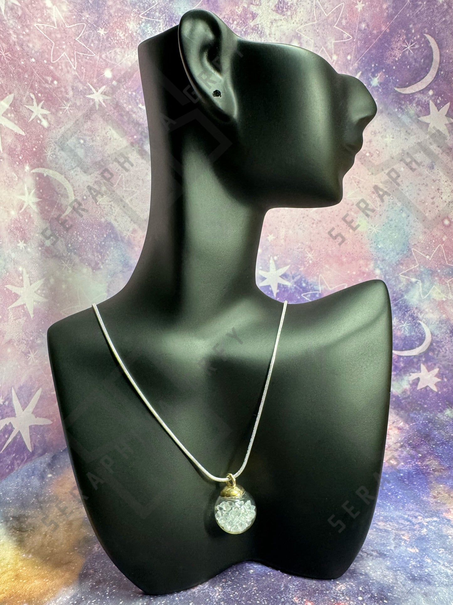 This image features a glass ball pendant necklace displayed on a black mannequin bust. The pendant is a glass ball with a gold-colored cap, filled with clear quartz crystals, hanging from a thin silver chain. The background is a colorful, galaxy-themed pattern with stars and nebula-like designs, enhancing the visual appeal of the necklace.