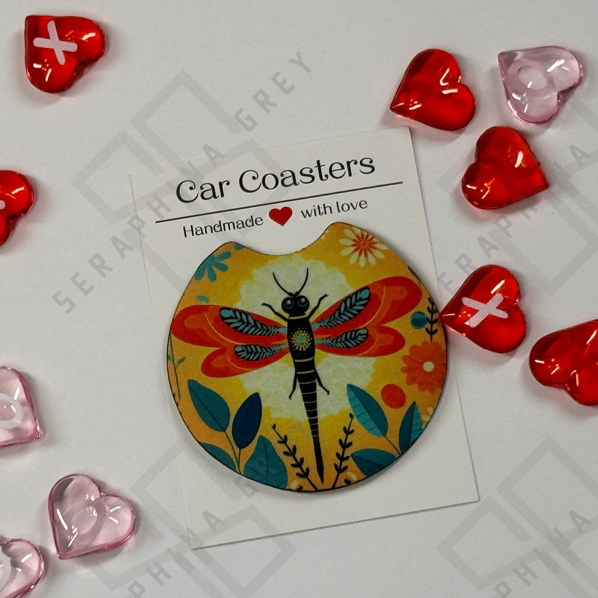 A single car coaster featuring a colorful design of a dragonfly with vibrant wings, set against a yellow floral background. The coaster is displayed with a label that reads ‘Car Coasters - Handmade with love.’ The background includes red and pink heart-shaped decorations