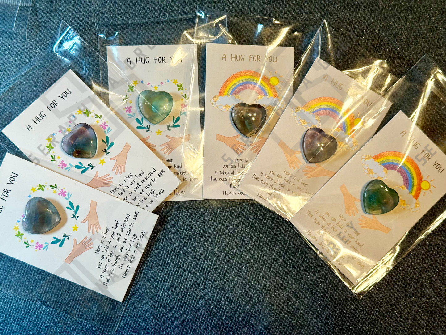 A collection of cards, each featuring a fluorite gemstone heart and the text ‘A Hug For You.’ The cards display different designs, including a floral wreath and a rainbow with clouds and sun. Each card has an illustration of two hands reaching out and includes a message conveying the warmth and comfort of a hug, even when apart. The cards are individually wrapped in plastic sleeves.