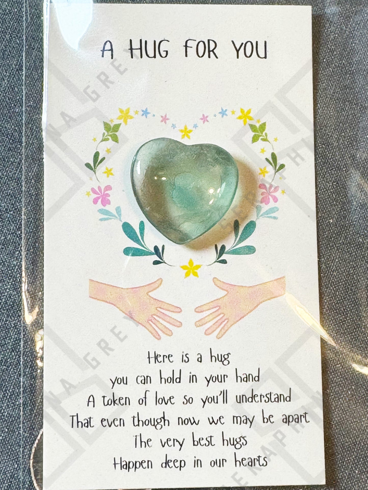 A card featuring a fluorite gemstone heart with the text ‘A Hug For You’ at the top. The design includes a floral wreath around the heart and an illustration of two hands reaching out. The message below the heart expresses that the heart stone is a token of love, offering comfort even when apart. The card is enclosed in a plastic sleeve.