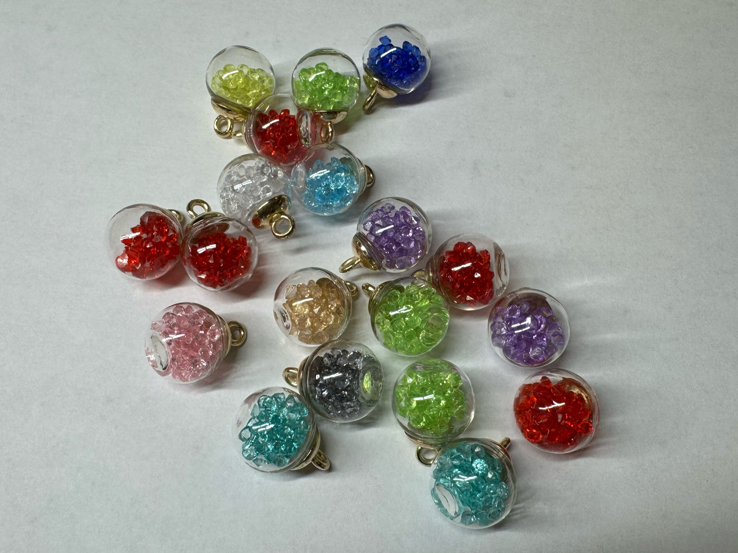 This image features a collection of glass globe pendants. Each pendant is a small clear glass sphere filled with colorful stones in various shades, including red, green, blue, purple, pink, and yellow. The pendants have gold-colored caps with loops for attaching to a necklace chain. They are arranged on a plain white background, highlighting the vibrant colors and intricate details of the pendants