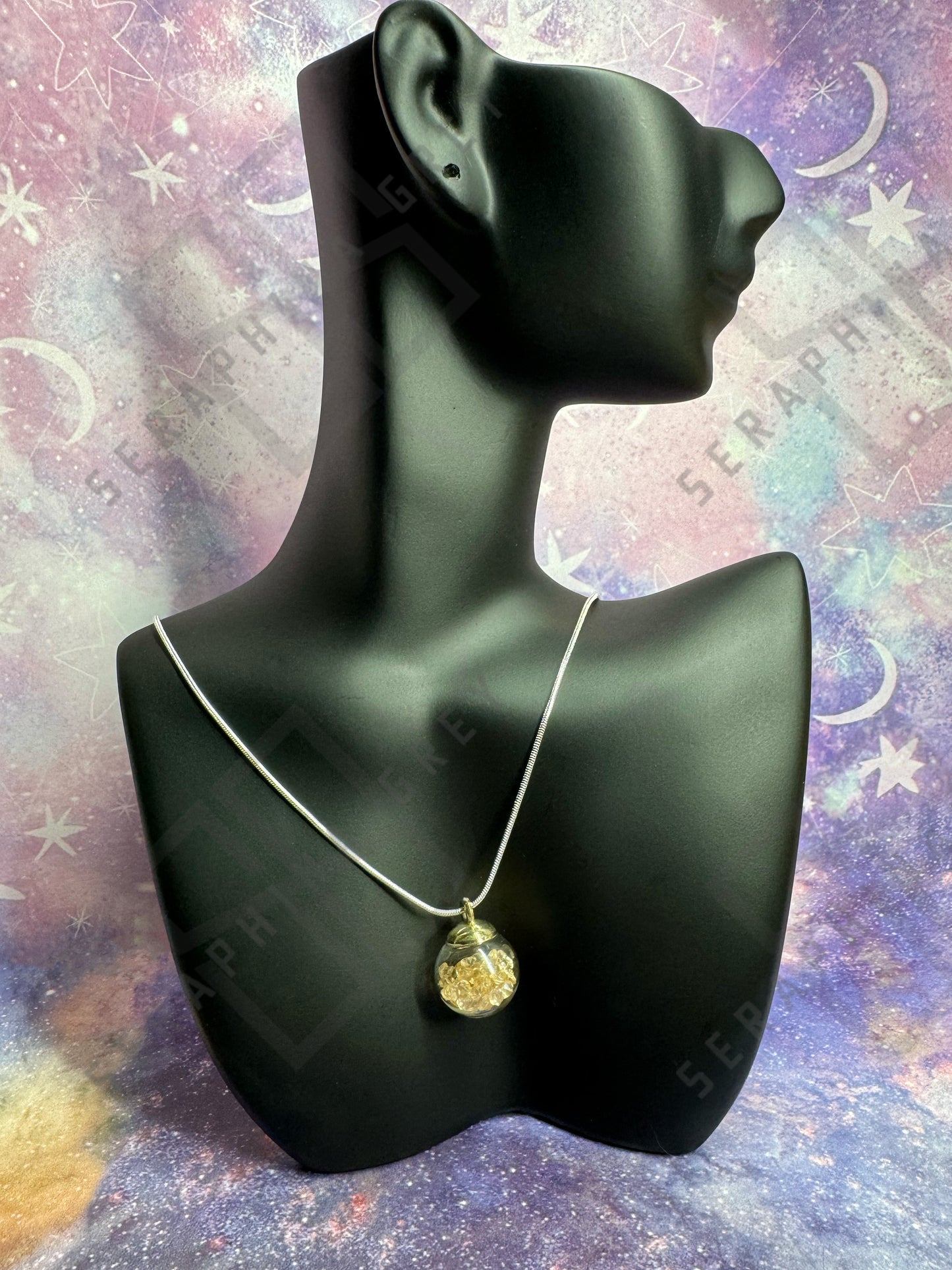 This image features a glass ball pendant necklace displayed on a black mannequin bust. The pendant is a glass ball with a gold-colored cap, filled with gold crystals, hanging from a thin silver chain. The background is a colorful, galaxy-themed pattern with stars and nebula-like designs, enhancing the visual appeal of the necklace.