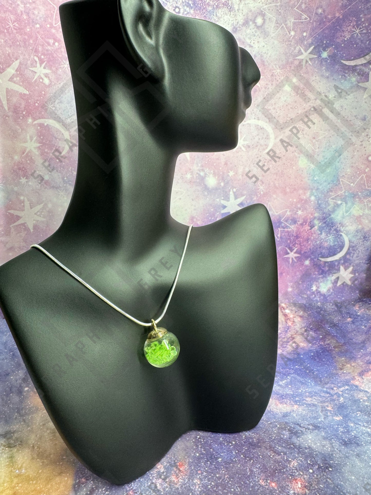 This image features a glass ball pendant necklace displayed on a black mannequin bust. The pendant is a glass ball with a gold-colored cap, filled with green crystals, hanging from a thin silver chain. The background is a colorful, galaxy-themed pattern with stars and nebula-like designs, enhancing the visual appeal of the necklace.