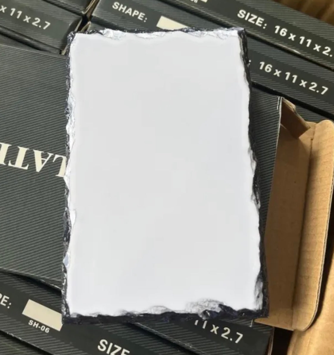 This image shows a custom photo slate with a blank white surface and a rectangular shape, featuring natural, uneven edges. The slate is placed on top of a stack of black boxes labeled with dimensions (“16 x 11 x 2.7”) and other product details. The background consists of these boxes, giving context to the slate’s packaging and storage. The watermark with “Seraphina Grey” is faintly visible across the background.