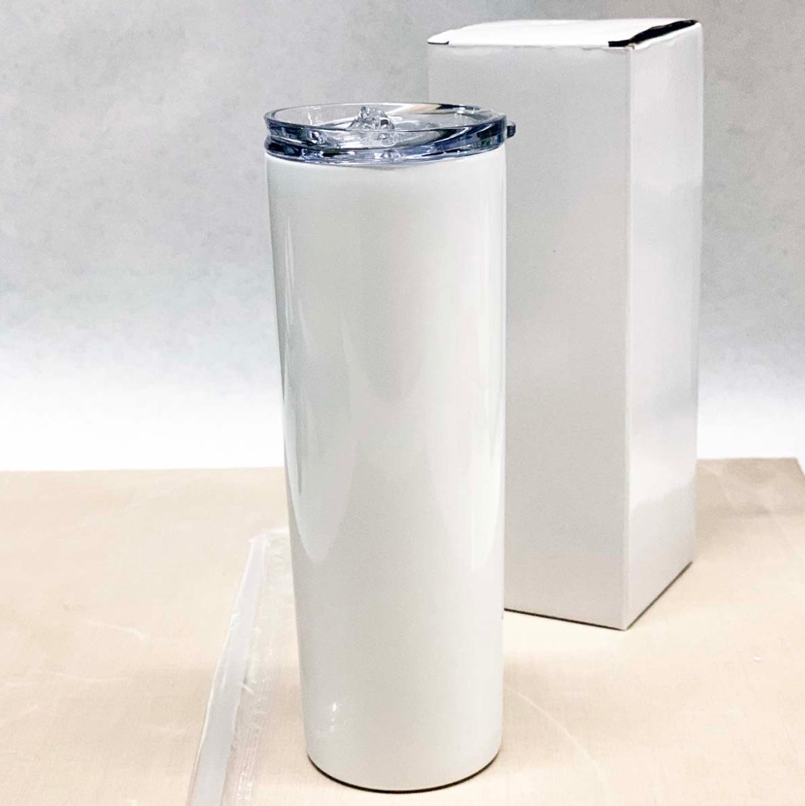 This image shows a sleek, white, 20 oz skinny tumbler with a clear lid. The tumbler is placed on a light-colored surface, and a white box is positioned behind it. The tumbler has a smooth and glossy finish, suitable for custom designs or logos.