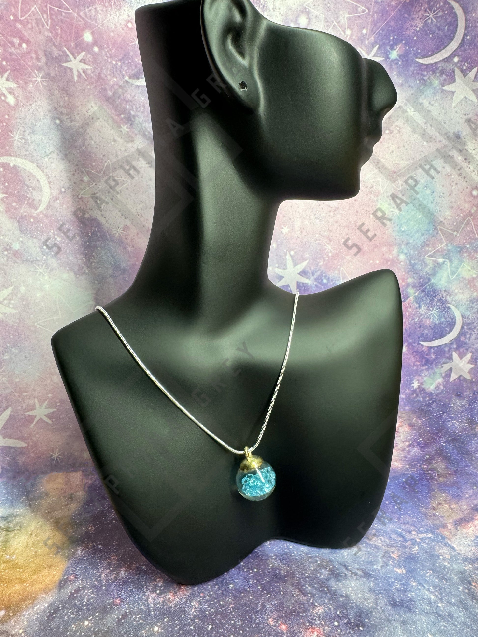 This image features a glass ball pendant necklace displayed on a black mannequin bust. The pendant is a glass ball with a gold-colored cap, filled with light blue crystals, hanging from a thin silver chain. The background is a colorful, galaxy-themed pattern with stars and nebula-like designs, enhancing the visual appeal of the necklace.