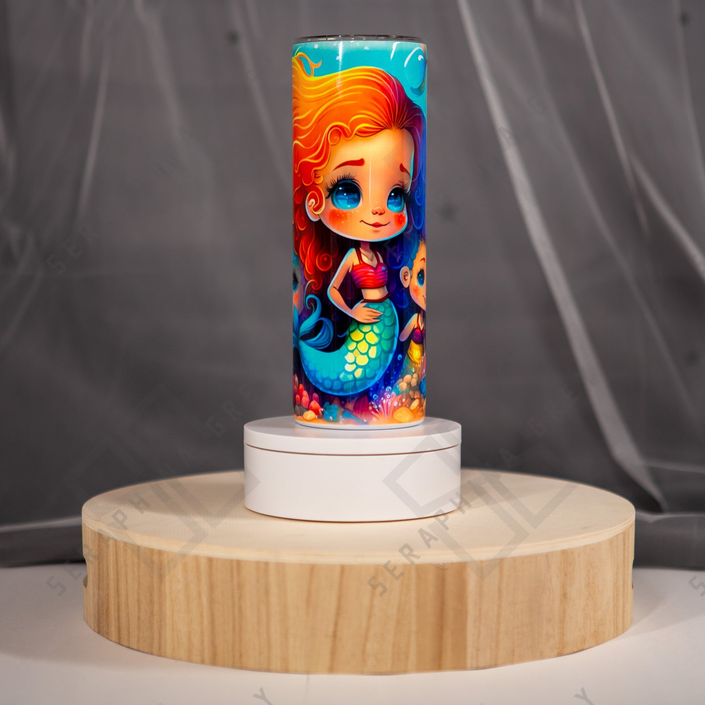 The image shows a colorful tumbler with a vibrant design featuring a cute mermaid. The mermaid has long, flowing red hair and is smiling, wearing a pink top and a green tail with blue scales. The background of the tumbler includes various underwater elements such as coral and bubbles in bright, cheerful colors. The tumbler is placed on a circular wooden stand with a white base, set against a gray background. The overall look is whimsical and appealing, ideal for anyone who loves mermaid-themed items.