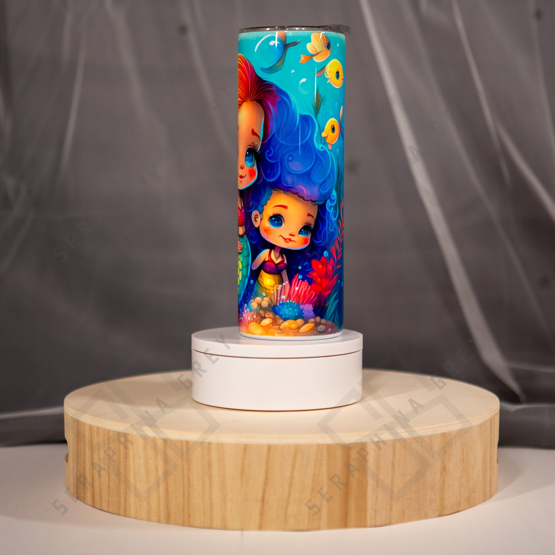 The image shows a side view of a vibrant tumbler design featuring a weemer with flowing blue hair beside her MerMom. They are surrounded by underwater elements such as coral, seaweed, bubbles and small fish near the top. The background is a bright blue, enhancing the underwater theme. The tumbler is placed on a circular wooden stand with a white base, set against a soft-focus gray background. The overall look is whimsical and appealing, ideal for anyone who loves mermaid-themed items.