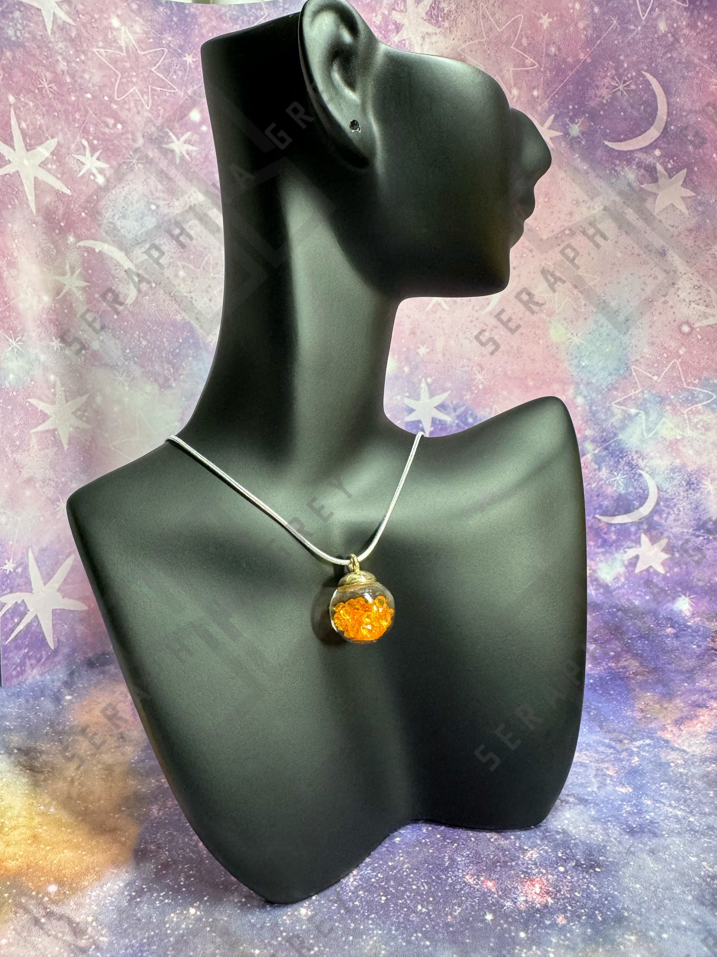 This image features a glass globe pendant necklace displayed on a black mannequin bust. The pendant is a glass ball with a gold-colored cap, filled with orange crystals, hanging from a thin silver chain. The background is a colorful, galaxy-themed pattern with stars and nebula-like designs, enhancing the visual appeal of the necklace.