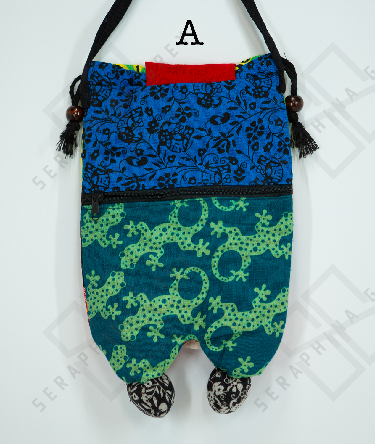 This image shows the backside of a handmade owl handbag labeled “A.” The back of the bag features two distinct fabric patterns: the upper part is a blue fabric with a black floral and bird design, and the lower part is a green fabric with a pattern of green lizards on a dark teal background. There is a black zipper separating the two fabric sections, adding functionality to the design. The bag has a black strap with wooden bead accents and tassels. It is displayed against a plain white background.