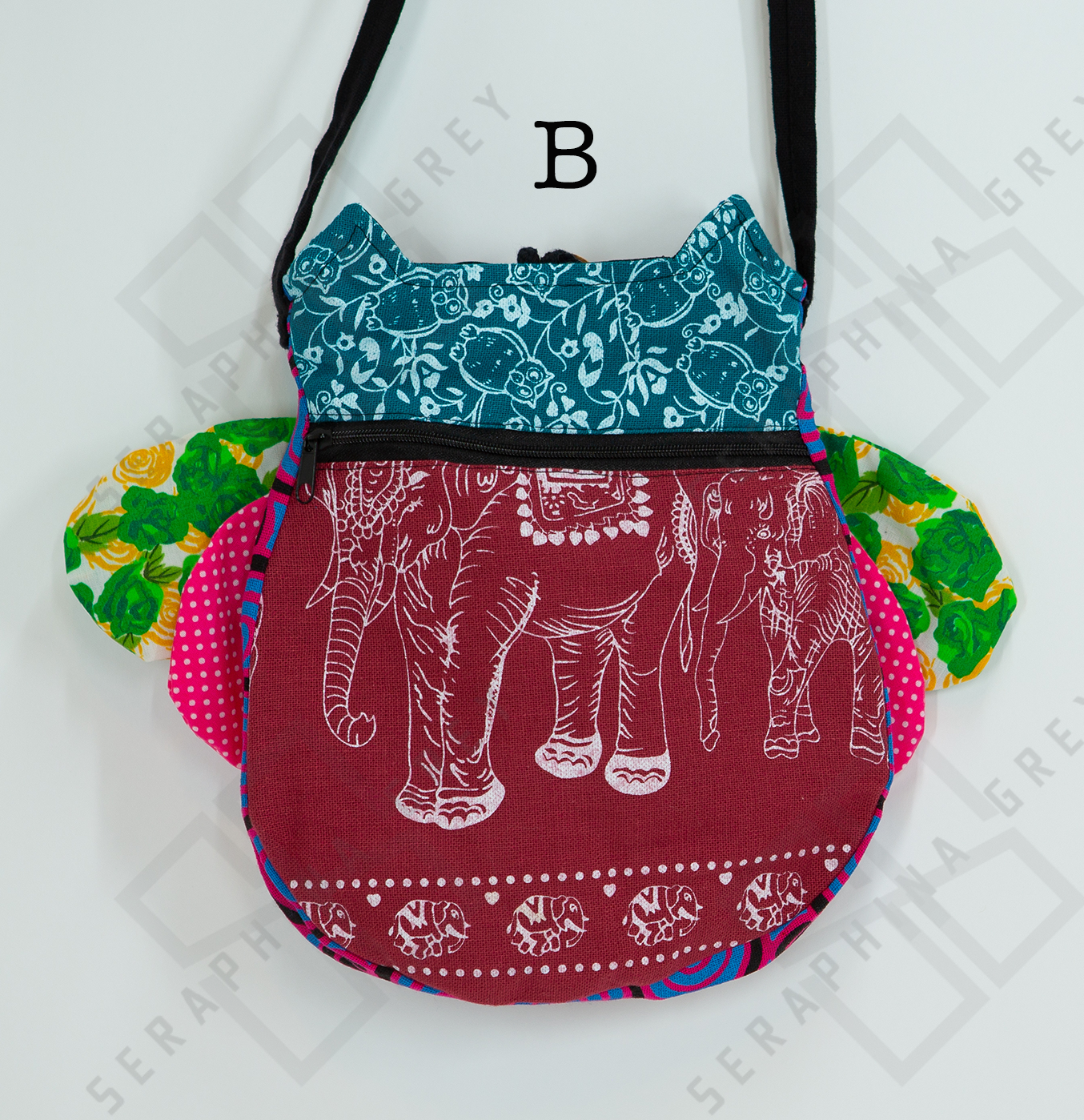 The image shows the back of a colorful handmade round owl handbag labeled “B.” The top half of the bag is blue-green with a white floral and owl pattern, while the bottom half is maroon with a white elephant design. The bag has a black zipper running horizontally across the middle. The owl’s green floral and pink polka-dotted wings are visible on the sides. The bag is hung by a black strap against a white background, highlighting its unique and playful design.