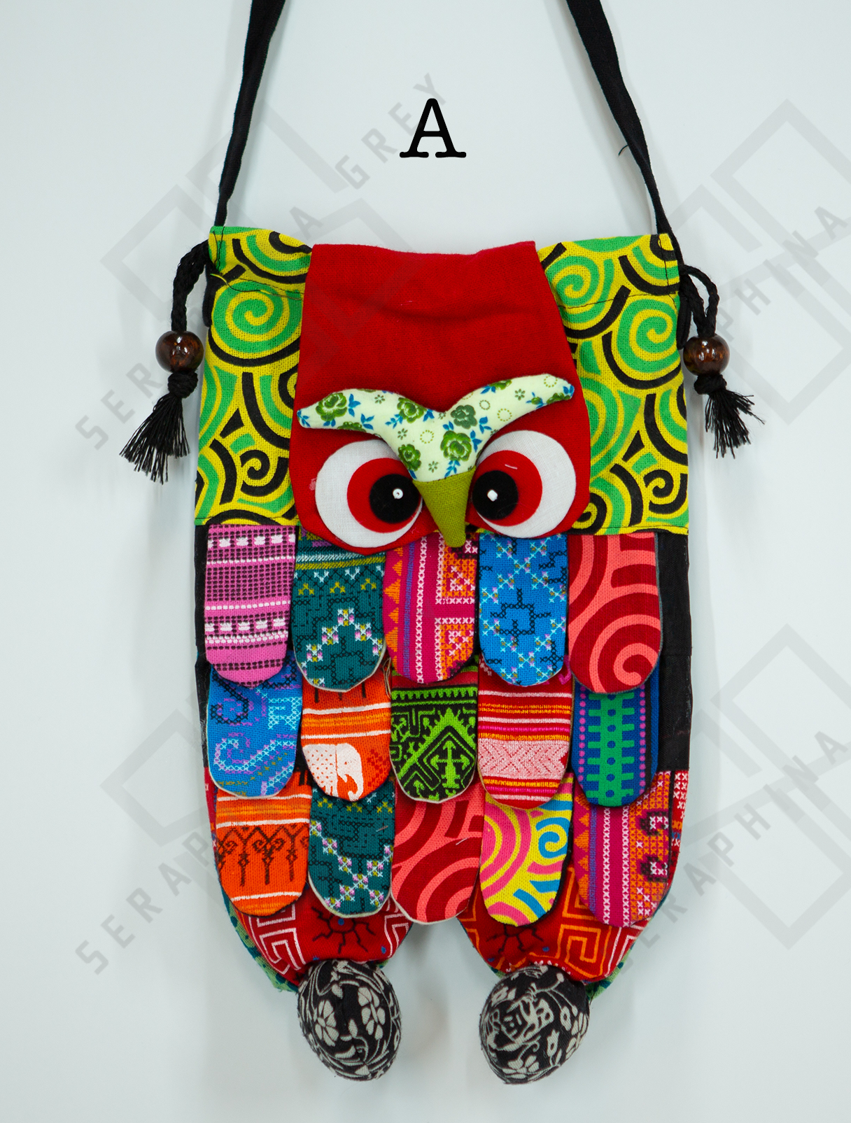 This image shows a single handmade owl handbag labeled “A.” The handbag features a colorful, patchwork design with a variety of vibrant patterns and fabrics. The owl face includes large, expressive eyes and a green beak set against a red background. The body of the owl is made up of multicolored fabric strips, giving it a feathered appearance. The bag has a black strap with wooden bead accents and tassels. It is displayed against a plain white background, clearly showcasing its intricate and playful design.