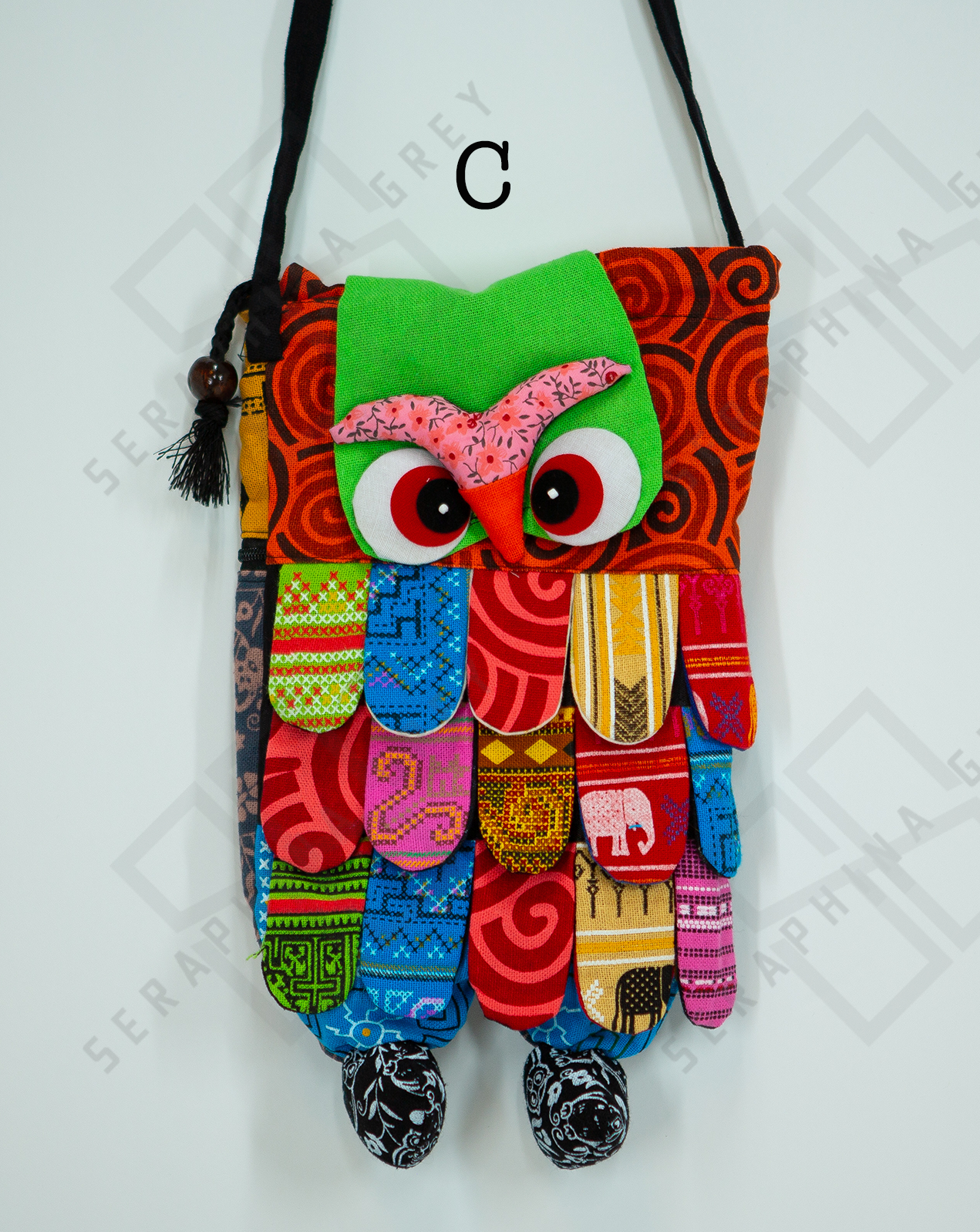 This image shows a single handmade owl handbag labeled “C.” The handbag features a colorful, patchwork design with various vibrant patterns and fabrics. The owl's face includes large, expressive eyes and a pink beak set against a green background. The body of the owl is made up of multicolored fabric strips, creating a feathered appearance. The bag has a black strap with wooden bead accents and tassels. It is displayed against a plain white background, clearly showcasing its intricate and playful design.