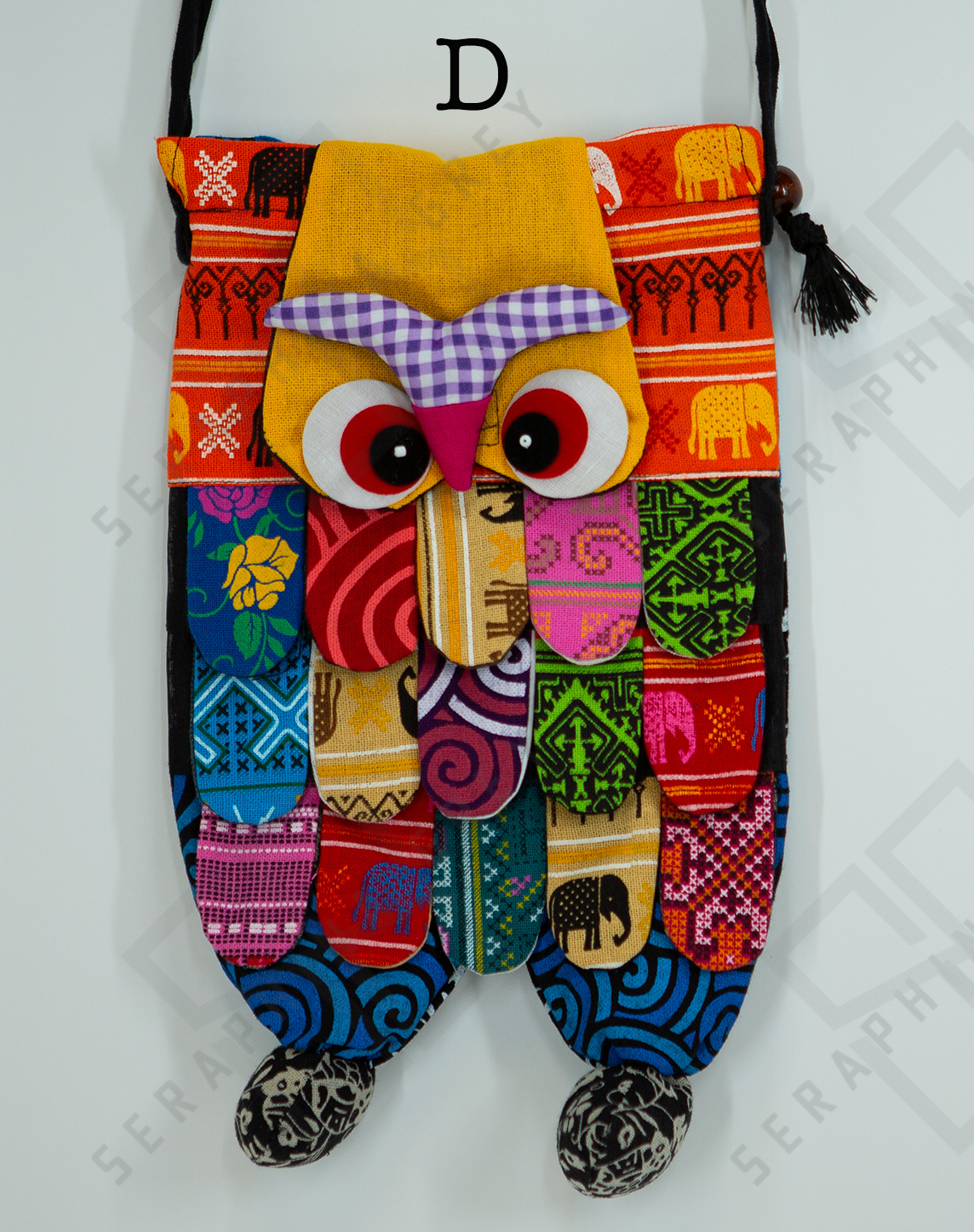 This image shows a single handmade owl handbag labeled “D.” The handbag features a colorful, patchwork design with a variety of vibrant patterns and fabrics. The owl’s face has large, expressive eyes and a pink beak set against a yellow background. The body of the owl is made up of multicolored fabric strips, giving it a feathered appearance. The bag has a black strap with wooden bead accents and tassels. It is displayed against a plain white background, clearly showcasing its intricate and playful design.