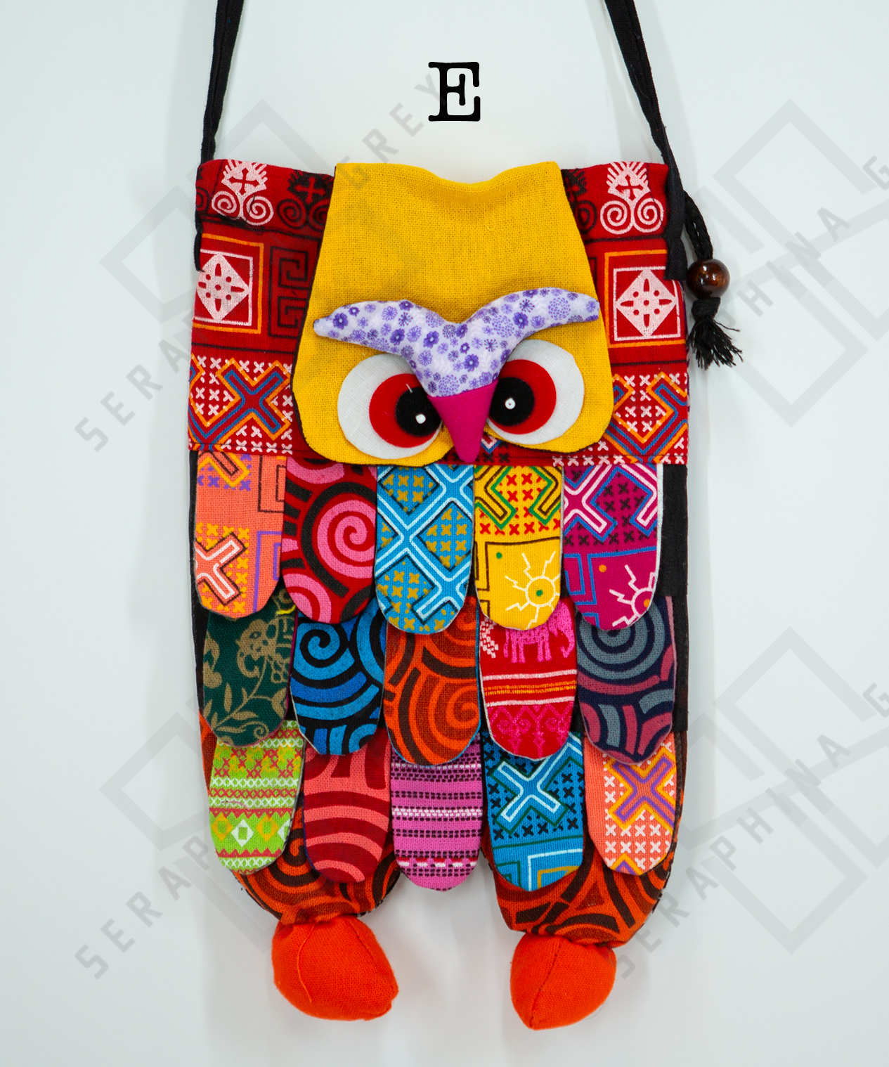 This image shows a single handmade owl handbag labeled “E.” The handbag features a colorful, patchwork design with various vibrant patterns and fabrics. The owl's face includes large, expressive eyes and a pink beak set against a yellow background. The body of the owl is made up of multicolored fabric strips, creating a feathered appearance. The bag has a black strap with wooden bead accents and tassels. It is displayed against a plain white background, clearly showcasing its intricate and playful design.