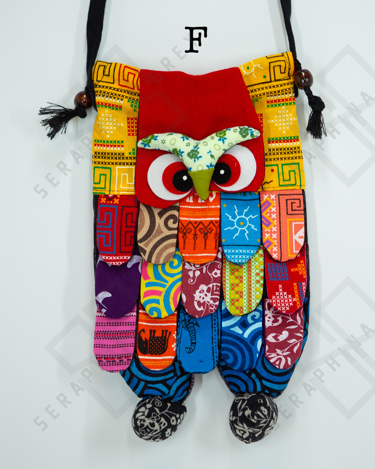 This image shows a single handmade owl handbag labeled “F.” The handbag features a colorful, patchwork design with various vibrant patterns and fabrics. The owl's face includes large, expressive eyes and a green beak set against a red background. The body of the owl is made up of multicolored fabric strips, creating a feathered appearance. The bag has a black strap with wooden bead accents and tassels. It is displayed against a plain white background, clearly showcasing its intricate and playful design.