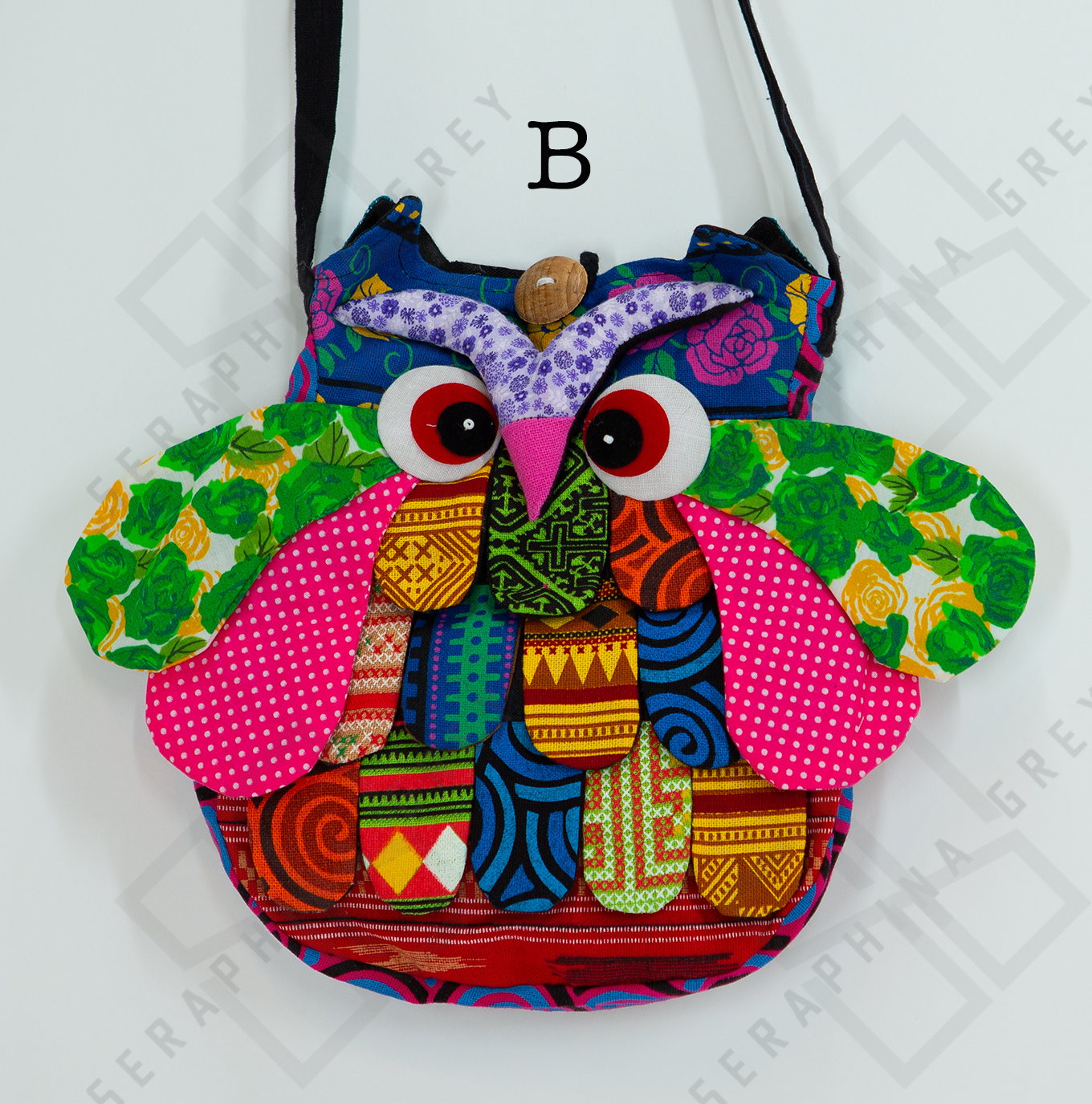 The image shows a colorful handmade round owl handbag labeled “B.” The bag features a vibrant patchwork design with various patterns, including green floral, pink polka dots, and geometric shapes in shades of yellow, red, blue, and orange. The owl’s large, expressive eyes and beak are prominently displayed, along with detailed wings and feathers. The bag is hung by a black strap against a white background, highlighting its whimsical and playful appearance.