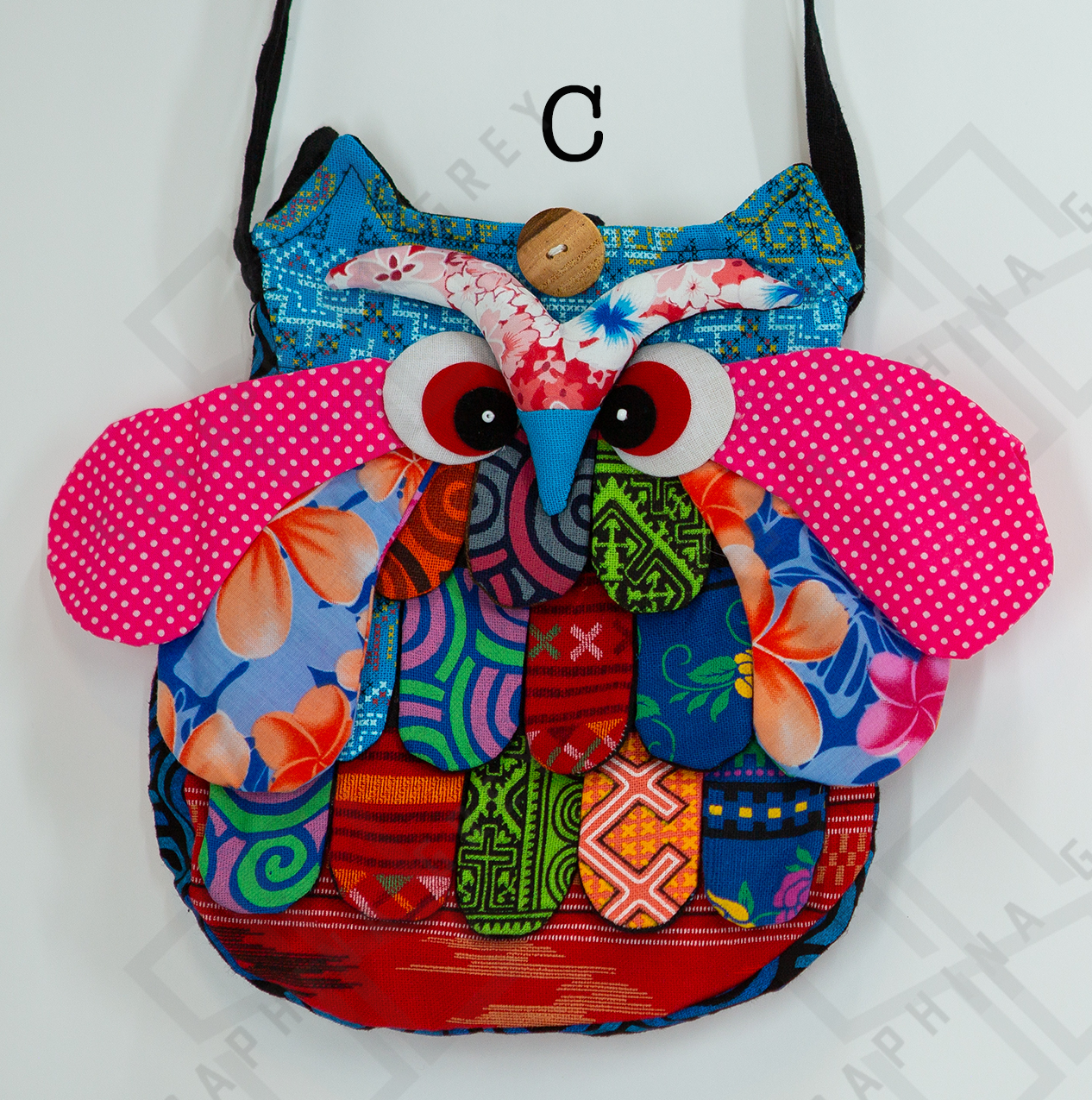 The image shows a colorful handmade round owl handbag labeled “C.” The bag features a vibrant patchwork design with various patterns, including floral, polka dots, and geometric shapes in shades of blue, pink, orange, green, and red. The owl’s large, expressive eyes and beak are prominently displayed, along with detailed wings and feathers. The bag is hung by a black strap against a white background, highlighting its whimsical and playful appearance