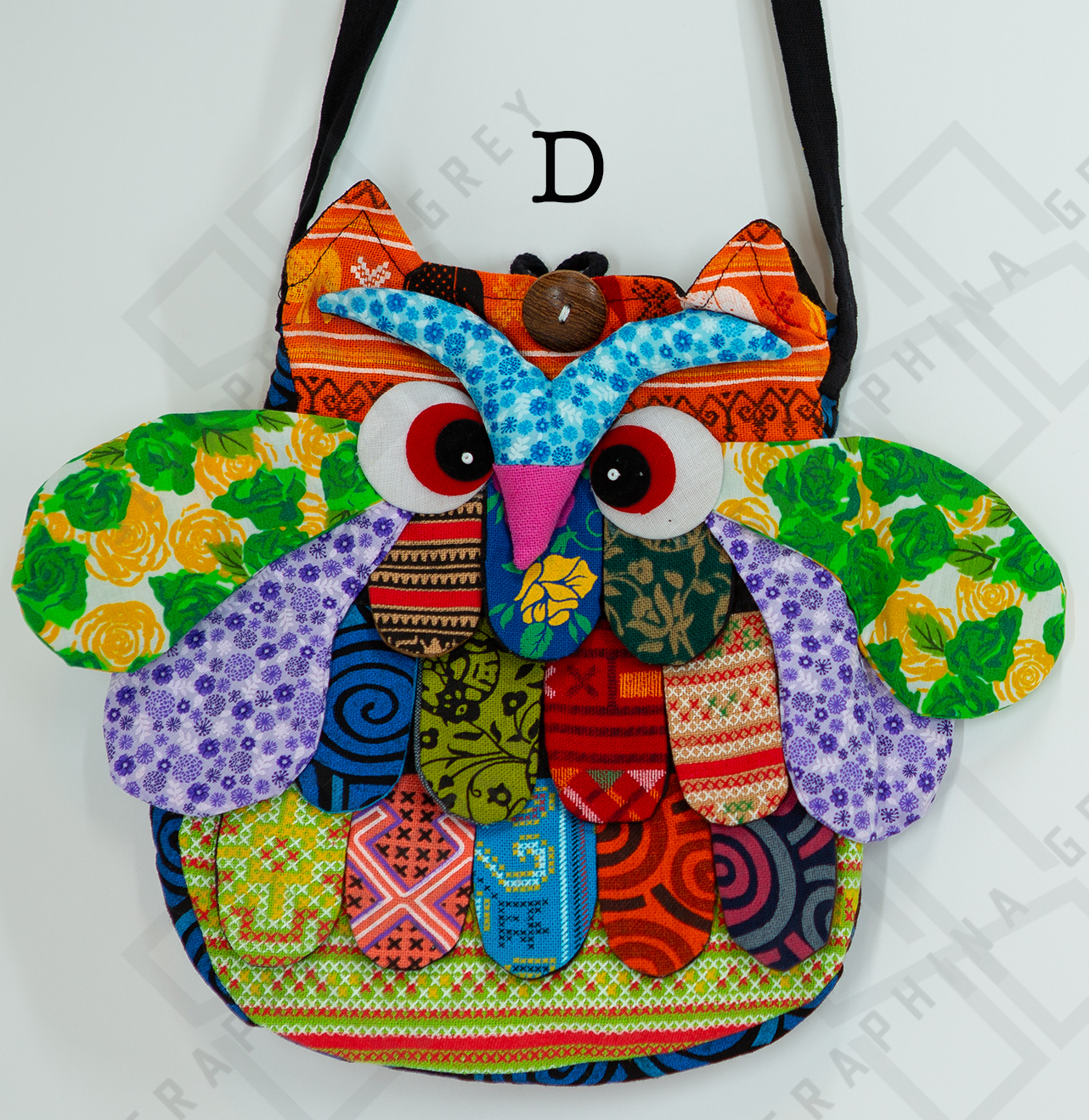 The image shows a colorful handmade round owl handbag labeled “D.” The bag features a vibrant patchwork design with various patterns, including floral, polka dots, and geometric shapes in shades of green, purple, yellow, blue, orange, and red. The owl’s large, expressive eyes and beak are prominently displayed, along with detailed wings and feathers. The bag is hung by a black strap against a white background, highlighting its whimsical and playful appearance.