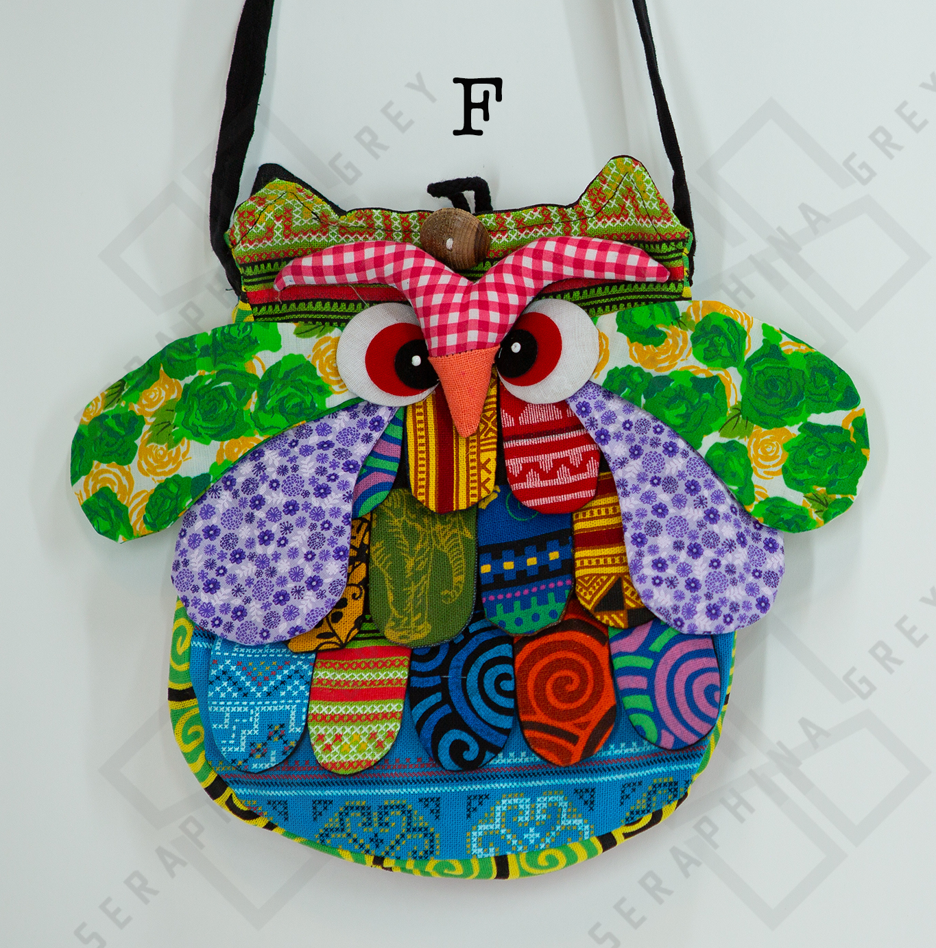 The image shows a colorful handmade round owl handbag labeled “F.” The bag features a vibrant patchwork design with various patterns, including green floral, purple polka dots, red gingham, and geometric shapes in shades of blue, yellow, and orange. The owl’s large, expressive eyes and beak are prominently displayed, along with detailed wings and feathers. The bag is hung by a black strap against a white background, highlighting its whimsical and playful appearance.