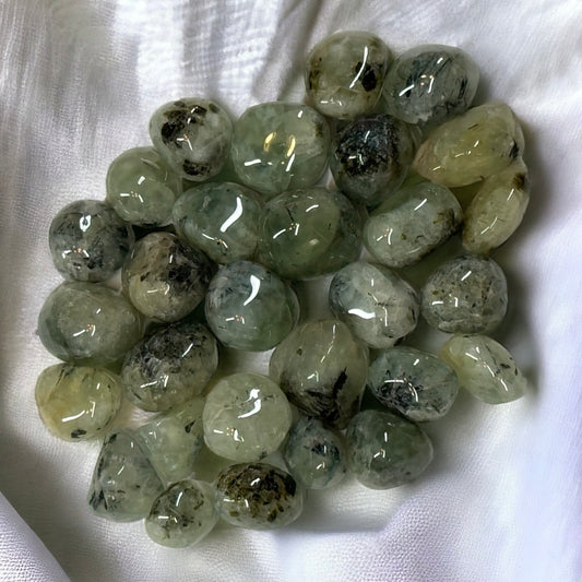 This image features a collection of Prehnite with Epidote gemstone pieces. The stones are polished and range in size from 0.75 to 1 inch. They have a translucent green color with dark inclusions of Epidote, giving them a unique and striking appearance. The gemstones are arranged on a white fabric background, highlighting their natural beauty and glossy finish.