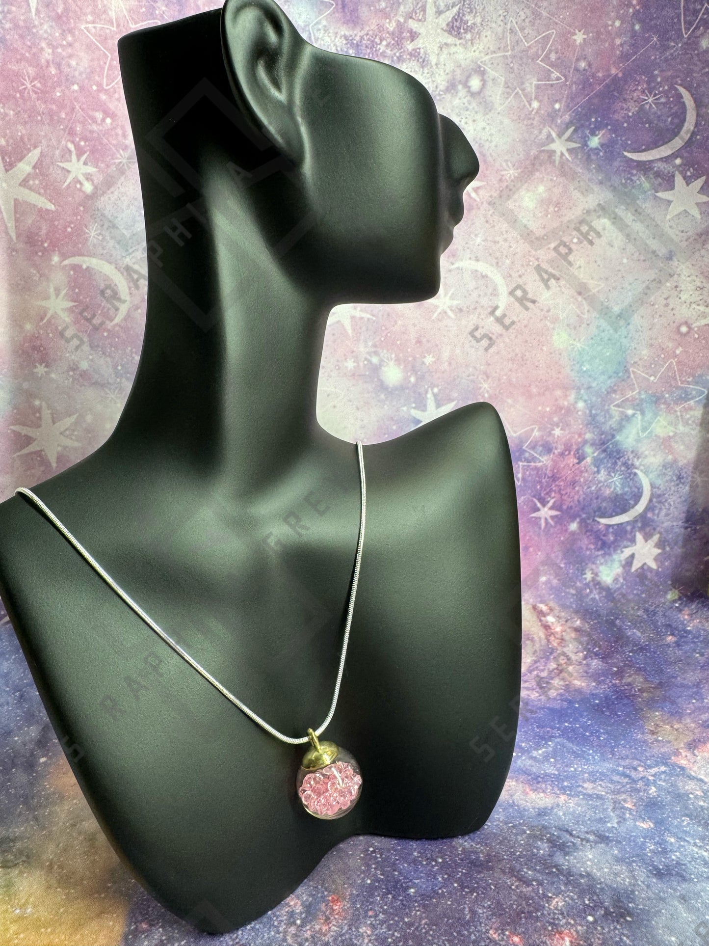 This image features a glass ball pendant necklace displayed on a black mannequin bust. The pendant is a glass ball with a gold-colored cap, filled with pink crystals, hanging from a thin silver chain. The background is a colorful, galaxy-themed pattern with stars and nebula-like designs, enhancing the visual appeal of the necklace.