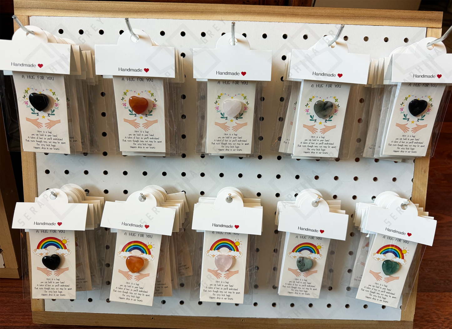 A market display featuring various ‘Pocket Hug’ cards hanging on a pegboard. Each card includes a gemstone heart and a heartfelt message, with some cards showcasing designs of floral wreaths or rainbows. The cards are individually packaged and labeled with ‘Handmade’ and a small red heart symbol. The display neatly arranges the different varieties, making them easily visible and accessible to shoppers.