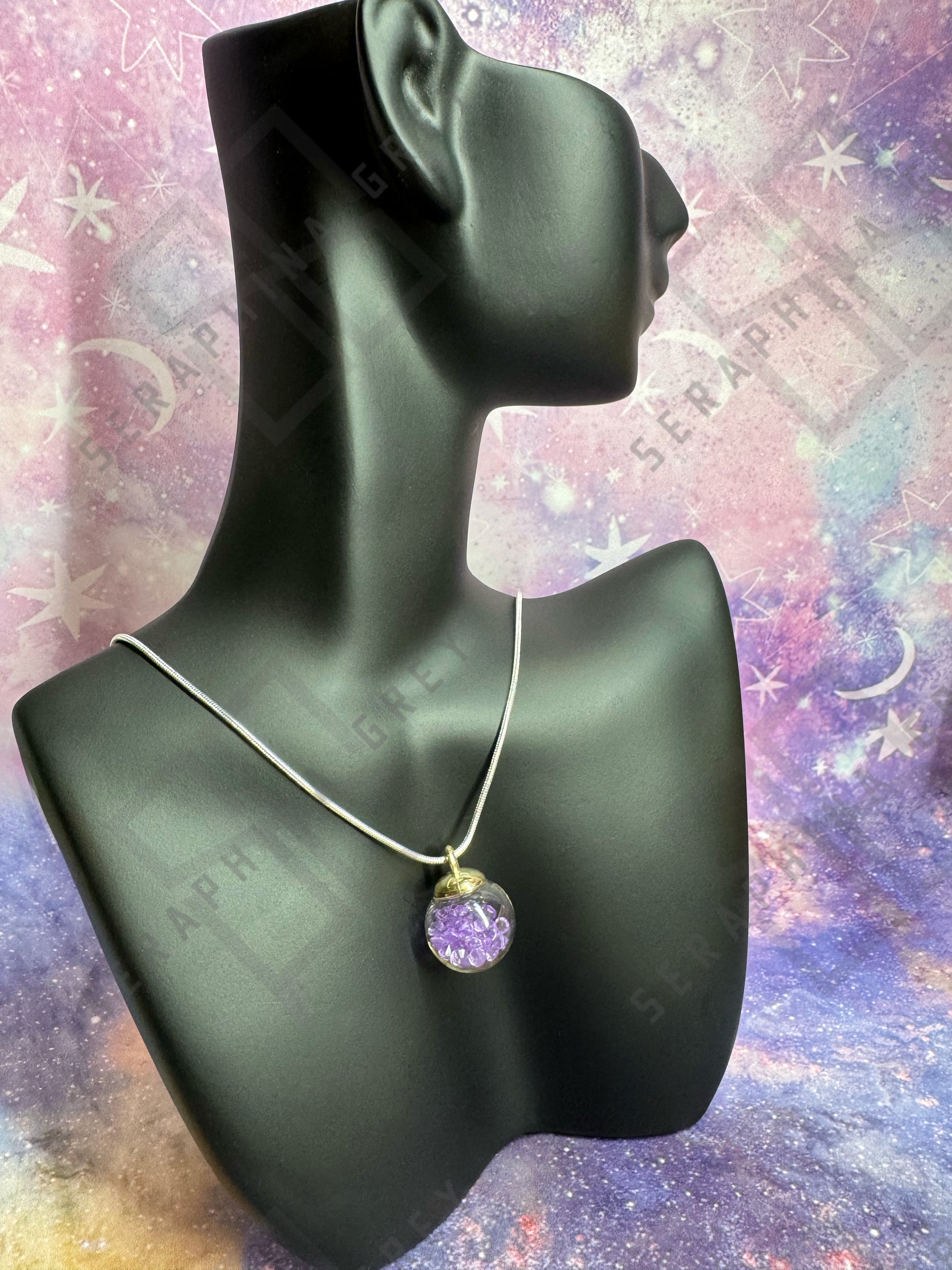 This image features a glass ball pendant necklace displayed on a black mannequin bust. The pendant is a glass ball with a gold-colored cap, filled with purple crystals, hanging from a thin silver chain. The background is a colorful, galaxy-themed pattern with stars and nebula-like designs, enhancing the visual appeal of the necklace.