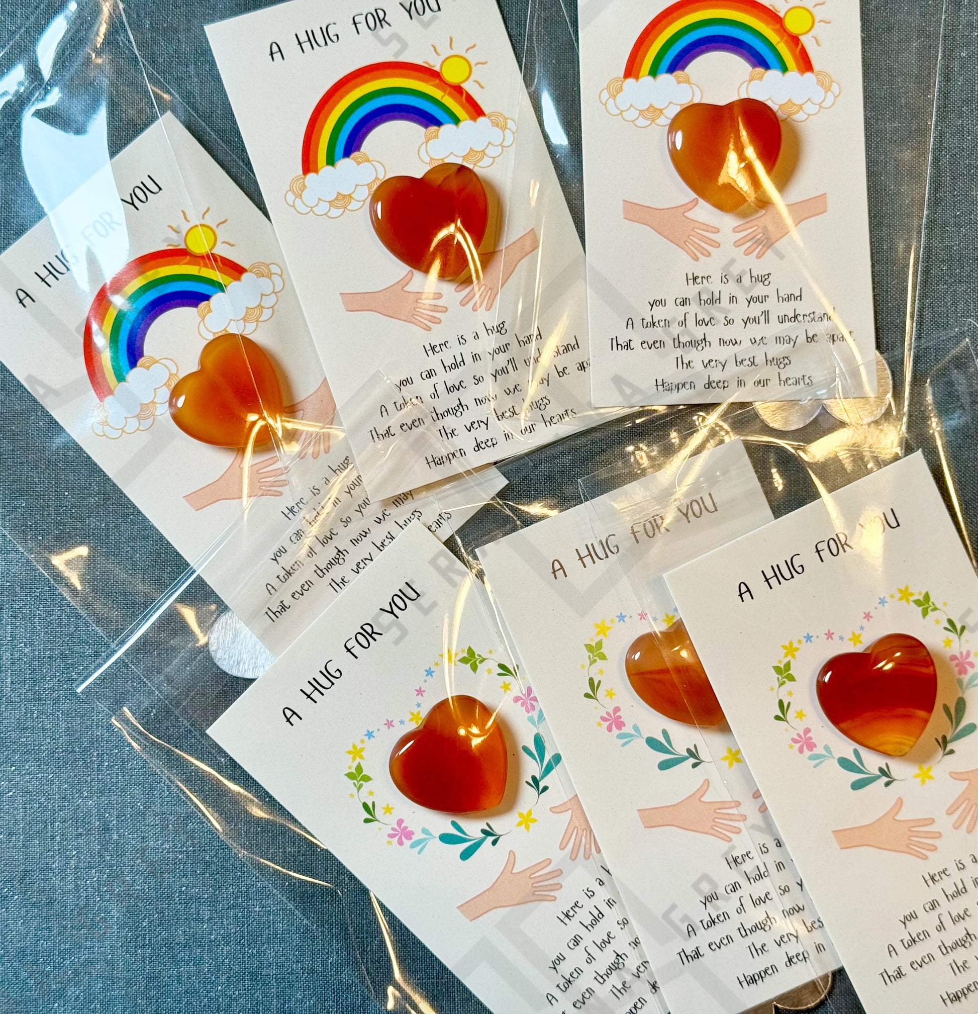 A collection of cards, each featuring a red carnelian heart and the text ‘A Hug For You.’ The cards showcase different designs, including a rainbow with clouds and sun, as well as a floral wreath. Each card has an illustration of two hands reaching out and a message expressing the warmth and comfort of a hug, even when separated. The cards are individually wrapped in plastic sleeves.