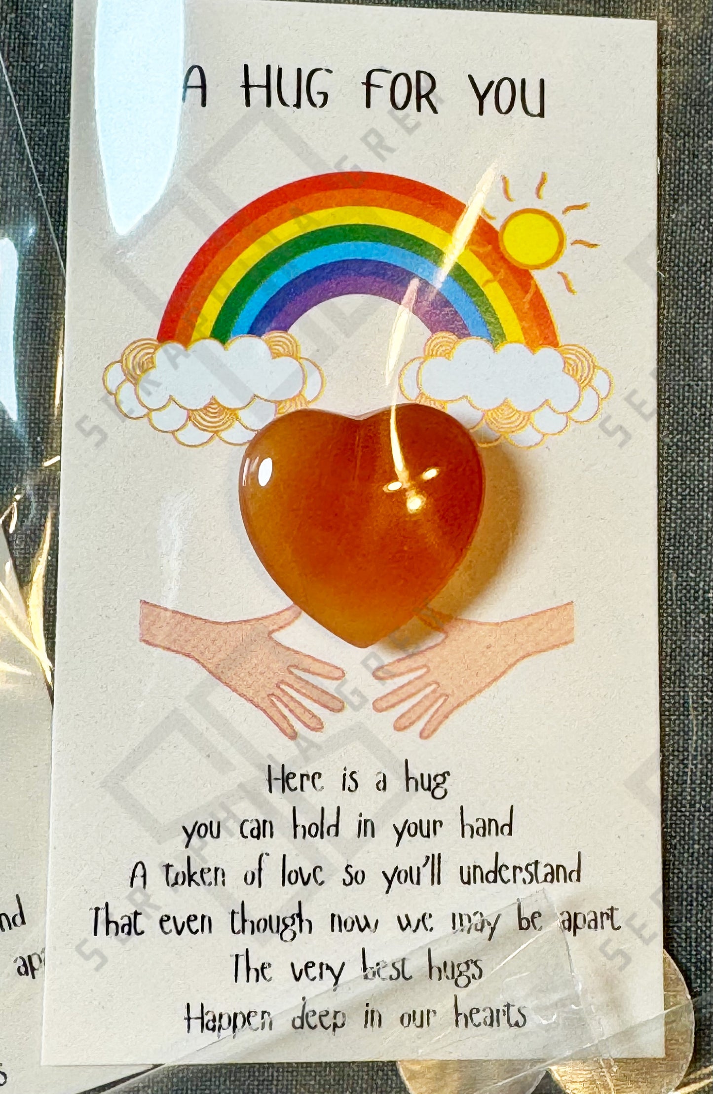 A card featuring a red carnelian heart with the text ‘A Hug For You’ at the top. The design includes a colorful rainbow with clouds and a shining sun above the heart, along with an illustration of two hands reaching out. The message below the heart expresses that the heart stone is a token of love, providing comfort even when apart. The card is wrapped in a plastic sleeve.