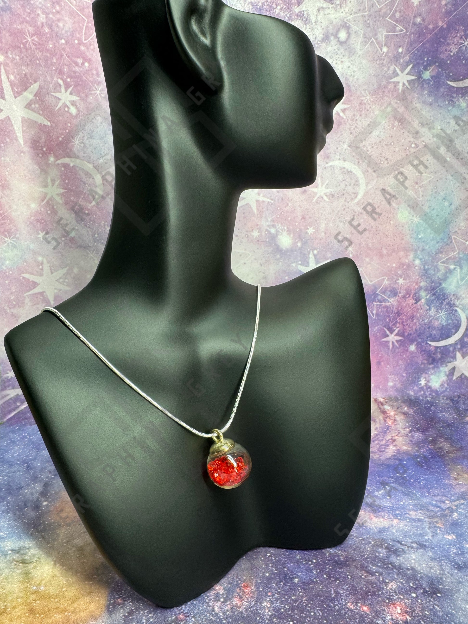 This image features a glass ball pendant necklace displayed on a black mannequin bust. The pendant is a glass ball with a gold-colored cap, filled with red crystals, hanging from a thin silver chain. The background is a colorful, galaxy-themed pattern with stars and nebula-like designs, enhancing the visual appeal of the necklace.