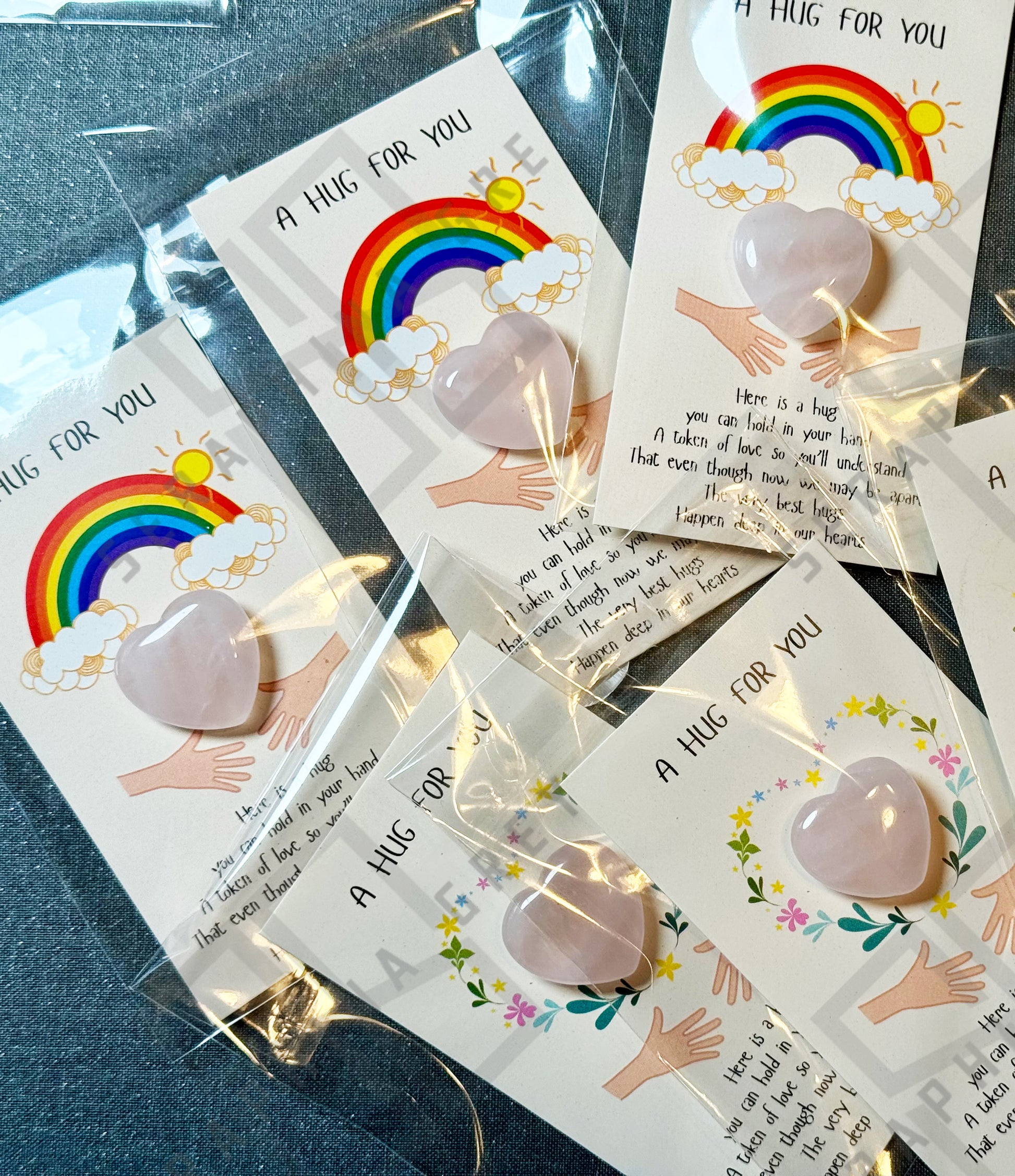 A collection of cards, each featuring a rose quartz heart and the text ‘A Hug For You.’ The cards include different designs, such as a colorful rainbow with clouds and sun, as well as a floral wreath. Each card has an illustration of two hands reaching out and a heartfelt message about the significance of hugs, despite being apart. The cards are individually packaged.