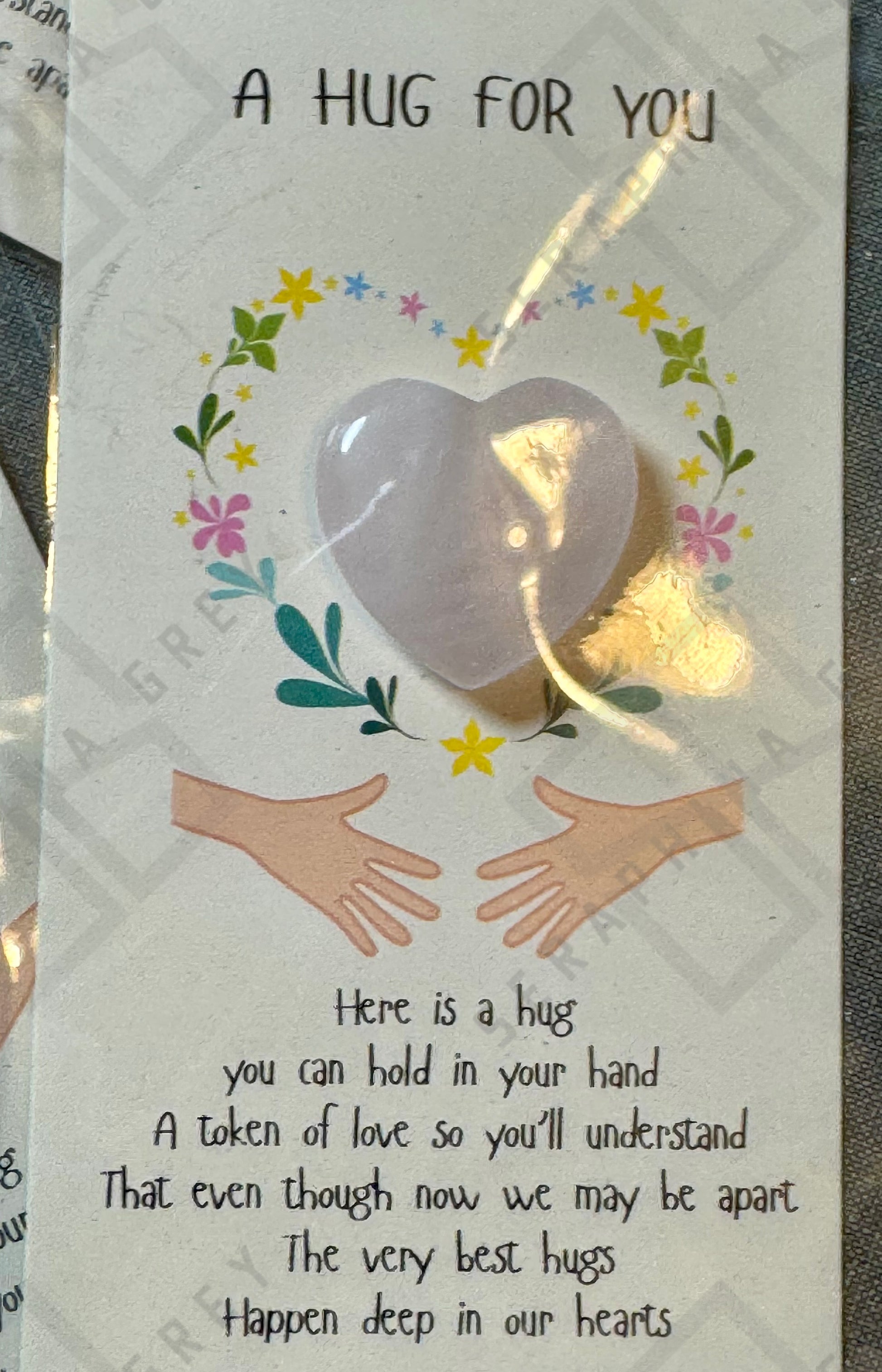 A card featuring a rose quartz heart with the text ‘A Hug For You’ at the top. The design includes an illustration of two hands reaching out with a floral wreath surrounding the heart. The text below reads: ‘Here is a hug you can hold in your hand, a token of love so you’ll understand that even though now we may be apart, the very best hugs happen deep in our heart