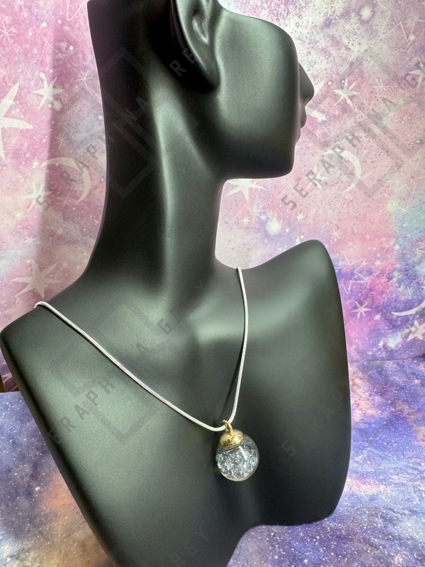 This image features a glass ball pendant necklace displayed on a black mannequin bust. The pendant is a glass ball with a gold-colored cap, filled with silver crystals, hanging from a thin silver chain. The background is a colorful, galaxy-themed pattern with stars and nebula-like designs, enhancing the visual appeal of the necklace.