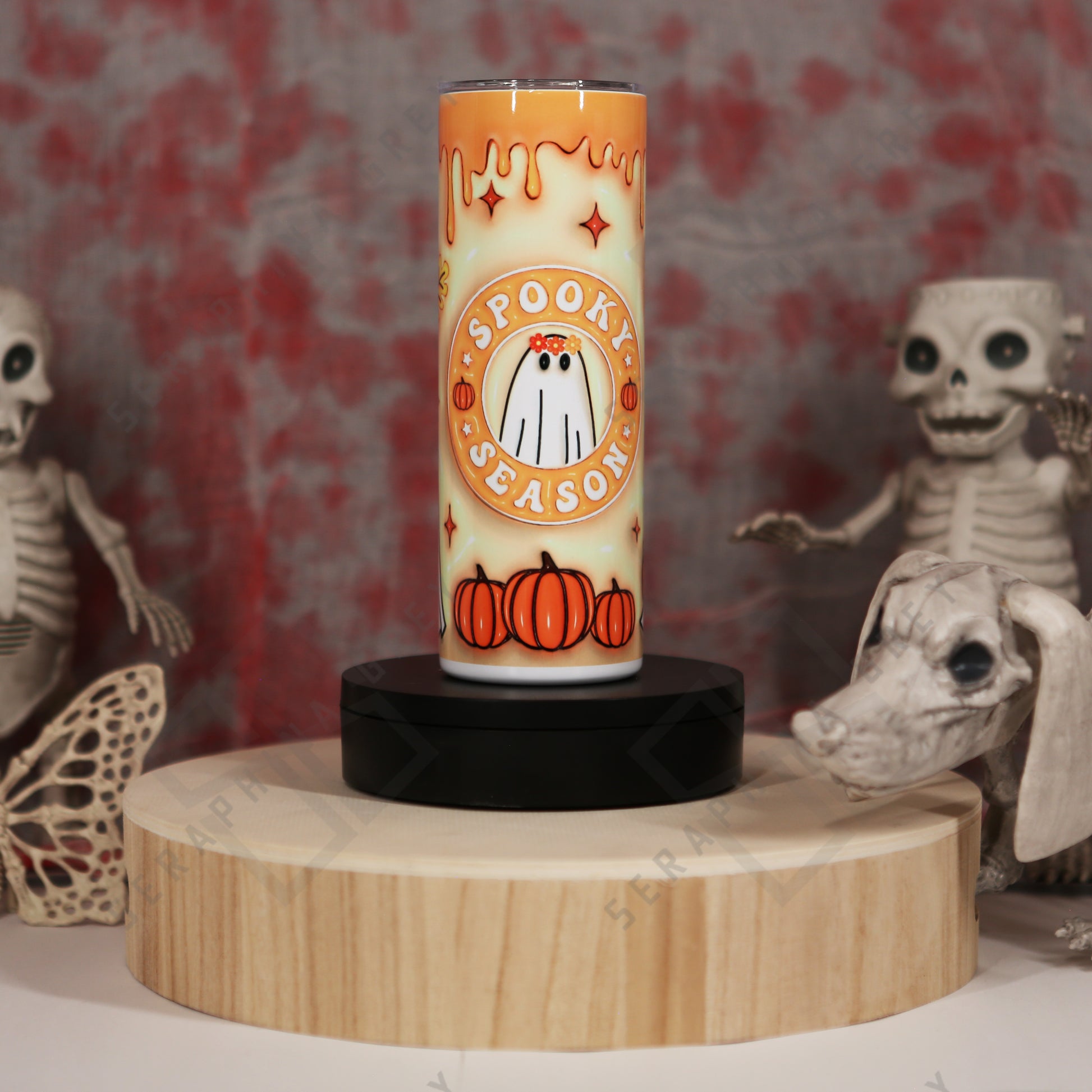 Orange drip Spooky Season autumn 20 oz tumbler. The tumbler features a gradient orange to cream background with dripping orange paint effect at the top. In the center, there is a cute ghost illustration with two small orange pumpkins at the bottom and the words ‘Spooky Season’ written around the ghost. The tumbler is placed on a round wooden platform, surrounded by Halloween-themed decorations, including skeletal figures and a skull in the background, enhancing the spooky ambiance.