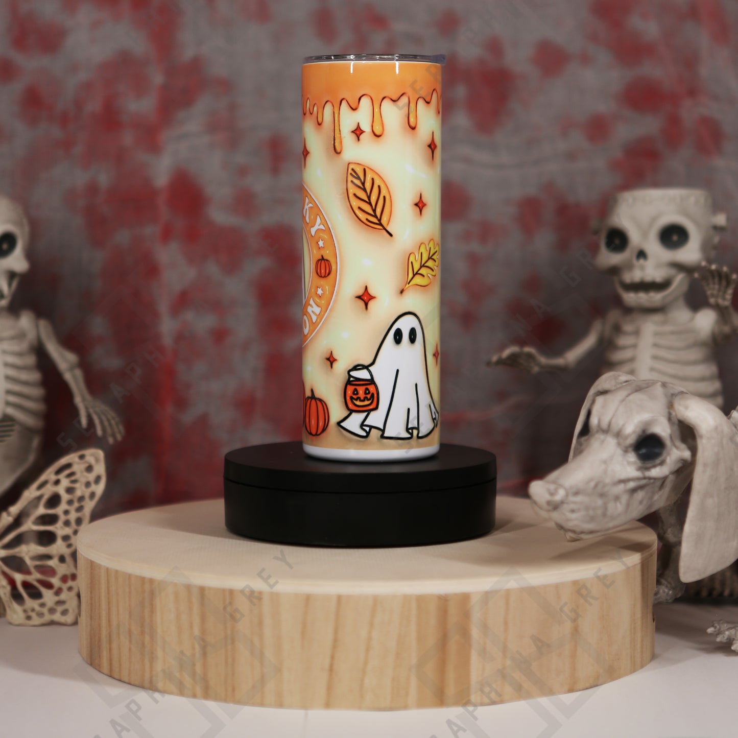 Orange drip Spooky Season autumn 20 oz tumbler. The tumbler features a gradient orange to cream background with a dripping orange paint effect at the top. It is decorated with autumn leaves, small star-like accents, and an illustration of a cute ghost holding a pumpkin bucket near the bottom. The tumbler is placed on a round wooden platform, surrounded by Halloween-themed decorations, including skeletal figures and a skull in the background, enhancing the spooky ambiance.