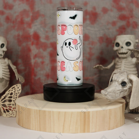 The image features a 20 oz tumbler with a colorful pastel 3D text that reads ‘Spooky Season,’ surrounding a cute ghost illustration holding a pink flower. Additional decorations include small black bats and crescent moons. The tumbler is placed on a round wooden platform, surrounded by Halloween-themed decorations, including skeletal figures in the background, enhancing the spooky and festive ambiance