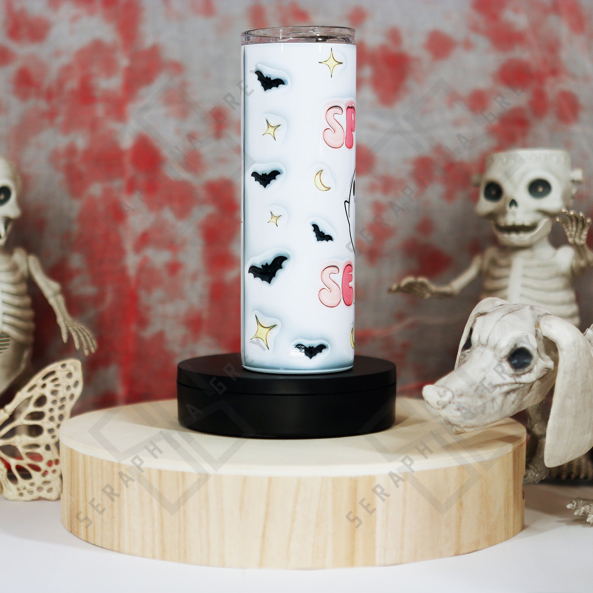 Side view of the Spooky Season 20 oz tumbler with a white background. The tumbler features colorful pastel text that reads ‘Spooky Season,’ surrounded by small black bats, crescent moons, and star-like accents. The tumbler is placed on a round wooden platform, surrounded by Halloween-themed decorations, including skeletal figures and a skull in the background, enhancing the spooky and festive ambiance.”