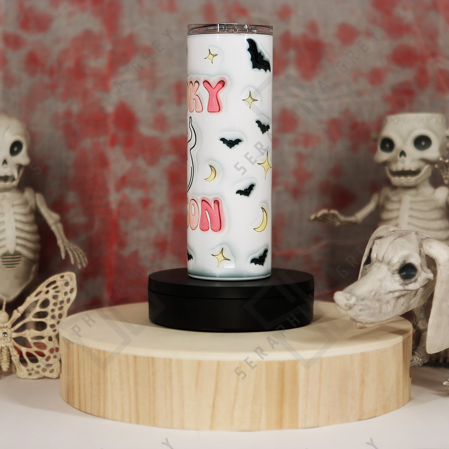 Side view of the Spooky Season 20 oz tumbler with a white background. The tumbler features colorful pastel text that reads ‘Spooky Season,’ surrounded by small black bats, crescent moons, and star-like accents. The tumbler is placed on a round wooden platform, surrounded by Halloween-themed decorations, including skeletal figures and a skull in the background, enhancing the spooky and festive ambiance.”