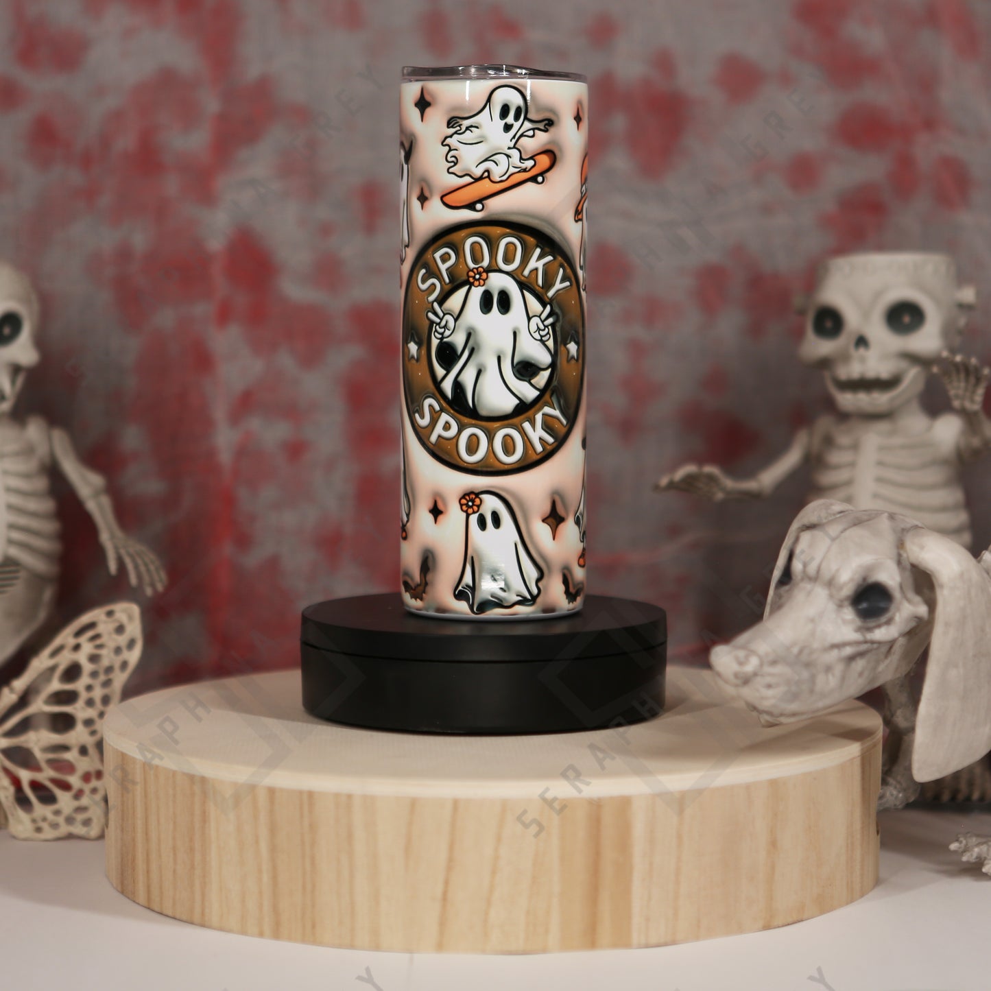 The image features a 20 oz tumbler with a beige and brown gradient background. The tumbler says Spooky Spooky" in the center with a cute ghost displaying the peace sign, in a circular pattern. Additional ghosts include a skater, a witch, trick or treater ghost, bats, and star-like accents scattered around the tumbler. The tumbler is placed on a round wooden platform, surrounded by Halloween-themed decorations, including skeletal figures in the background, enhancing the spooky ambiance.