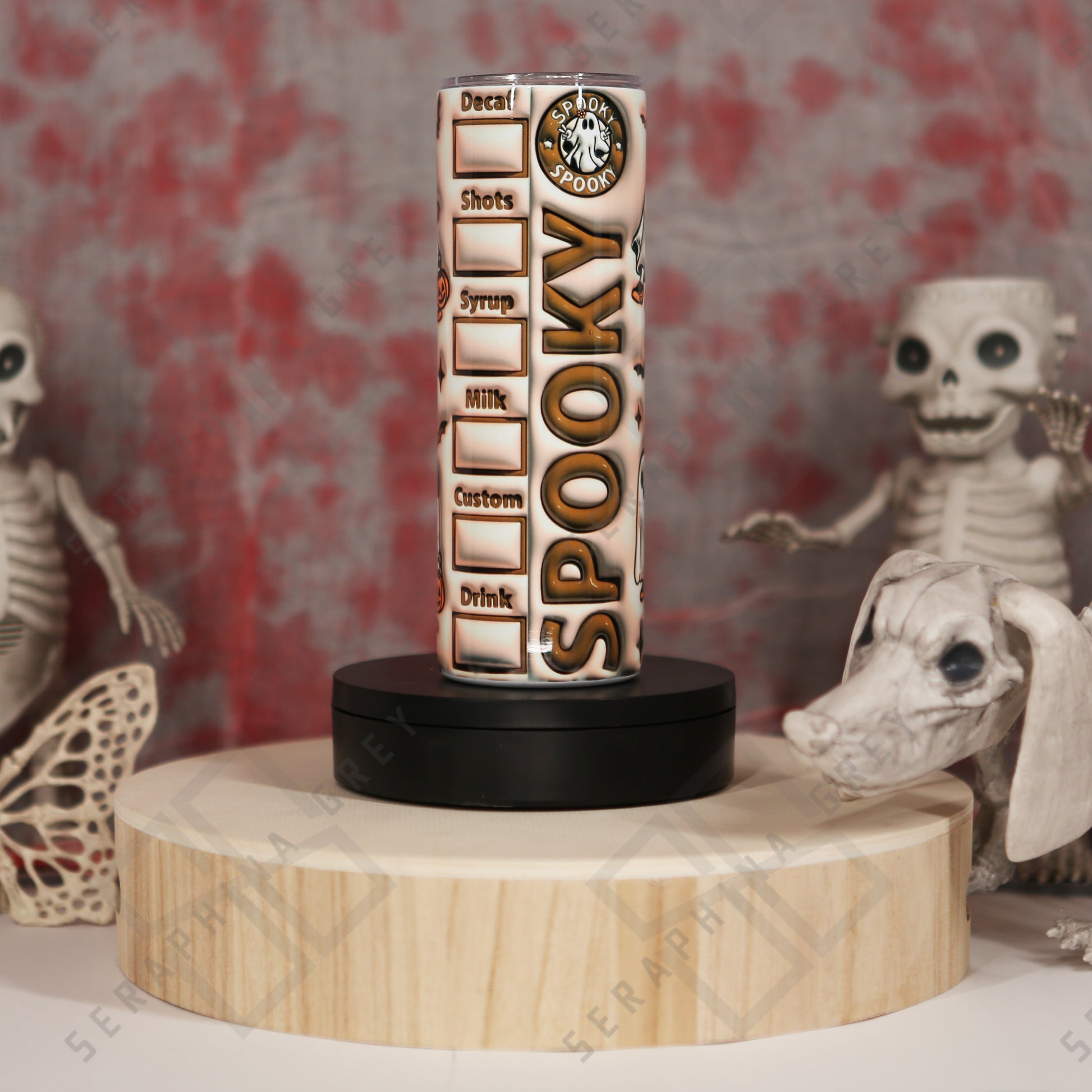 The image includes a 20 oz tumbler with ‘Spooky’ written vertically in large letters. It has a coffee order checklist design with checkboxes for decaf, shots, syrup, milk, custom, and drink. The tumbler is decorated with a ghost holding a pumpkin bucket and surrounded by small bats. It’s placed on a wooden platform with Halloween decorations, including skeletal figures in the background.