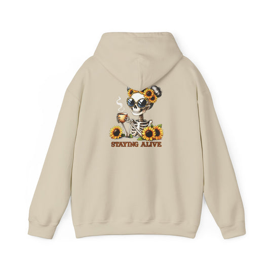 Sand-colored pullover hoodie shown from the back, featuring a skeleton wearing oversized sunglasses and sunflower space buns, holding a steaming mug, with “STAYING ALIVE” printed below.