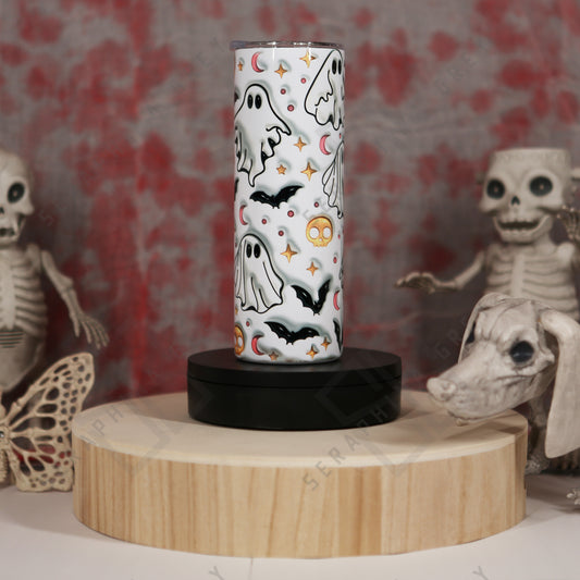 This image features a 20 oz skinny tumbler with a Halloween-themed design. The tumbler is decorated with 3D puffy ghosts, bats, crescent moons, and stars, all in a white background. The tumbler is displayed on a round wooden stand with a black base. The background includes Halloween decorations such as skeleton figures and a skull, adding to the spooky theme