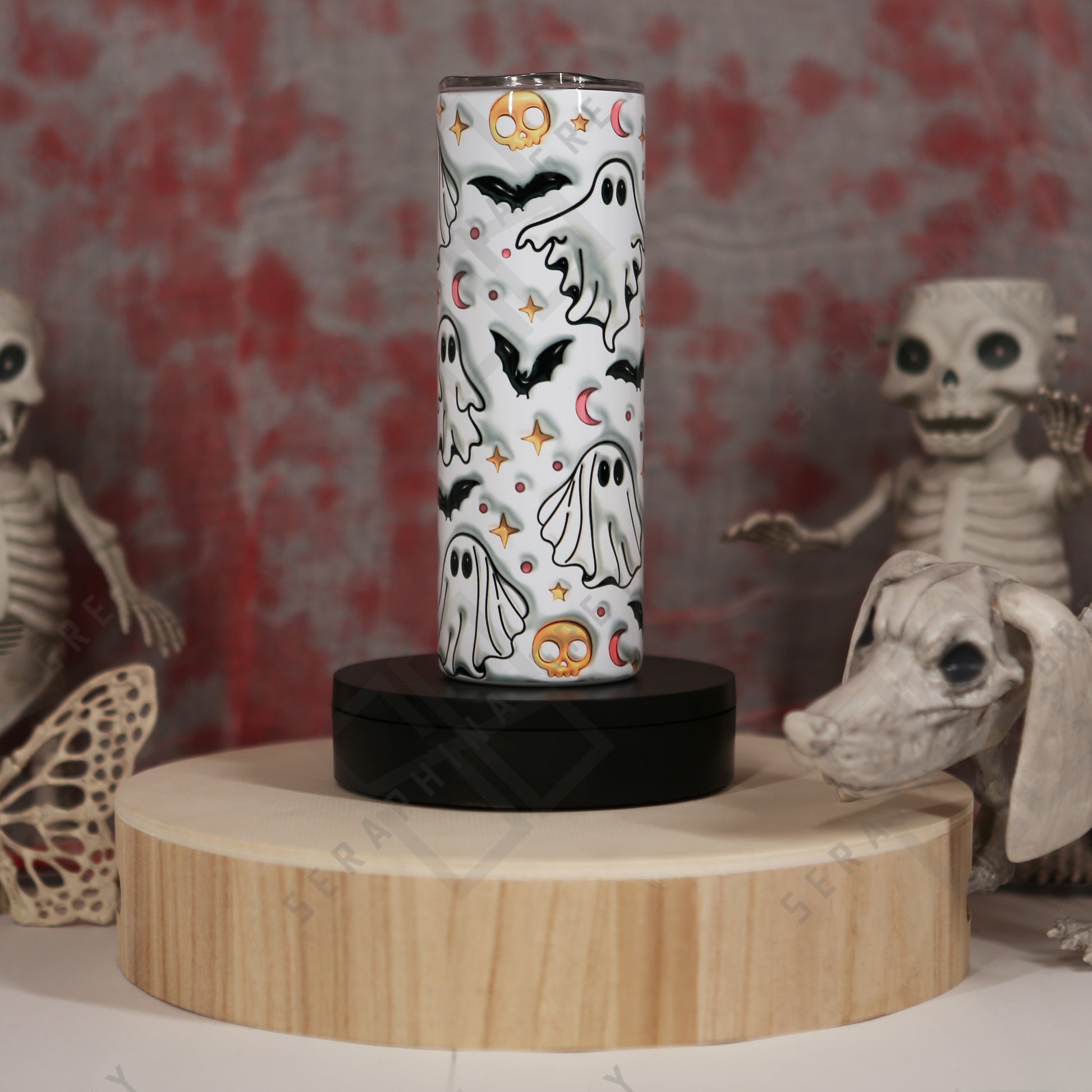 This image features a 20 oz skinny tumbler with a Halloween-themed design. The tumbler is decorated with 3D puffy ghosts, bats, crescent moons, and stars, all in a white background. The tumbler is displayed on a round wooden stand with a black base. The background includes Halloween decorations such as skeleton figures and a skull, adding to the spooky theme