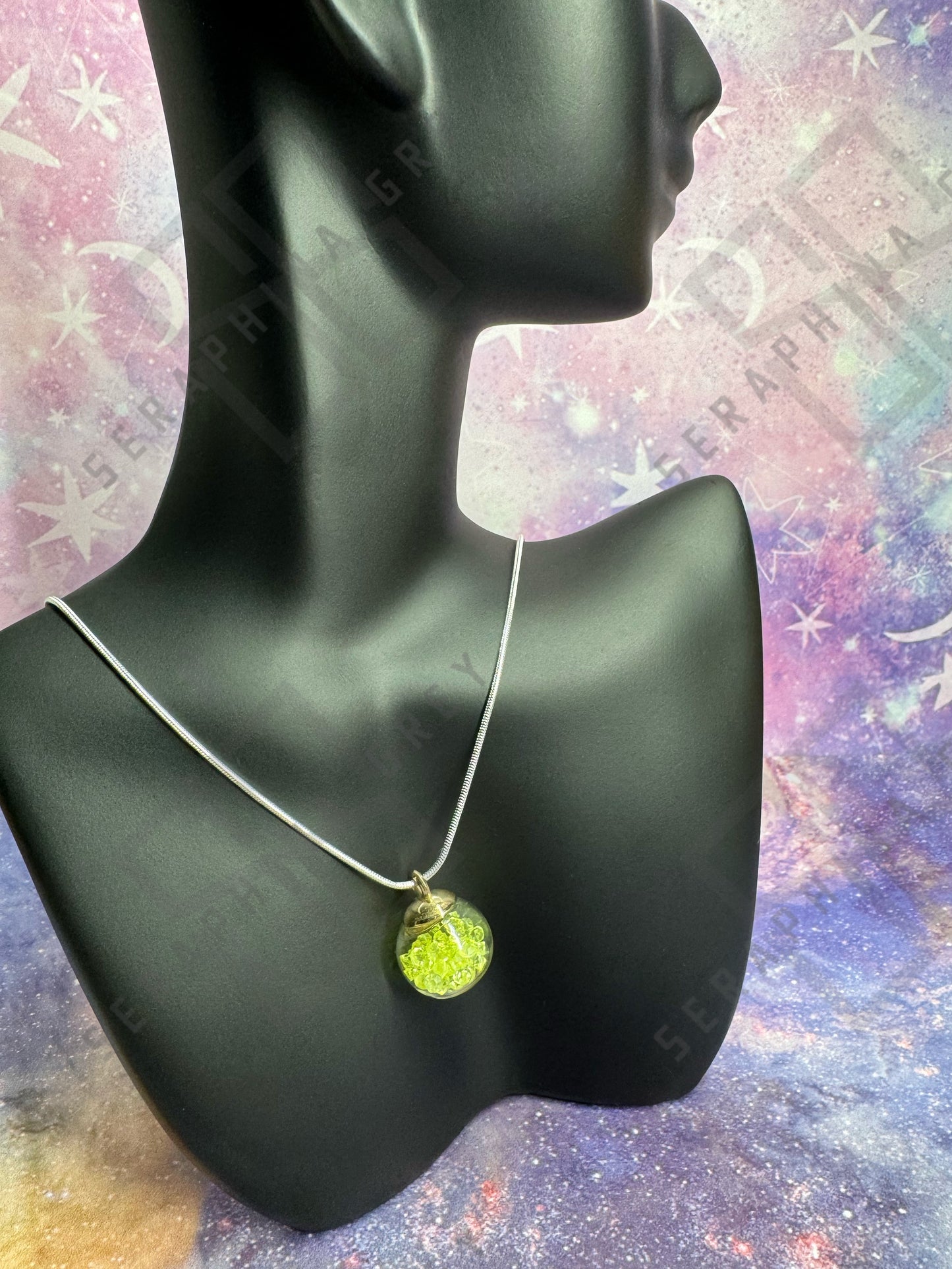 This image features a glass ball pendant necklace displayed on a black mannequin bust. The pendant is a glass ball with a gold-colored cap, filled with yellow crystals, hanging from a thin silver chain. The background is a colorful, galaxy-themed pattern with stars and nebula-like designs, enhancing the visual appeal of the necklace.