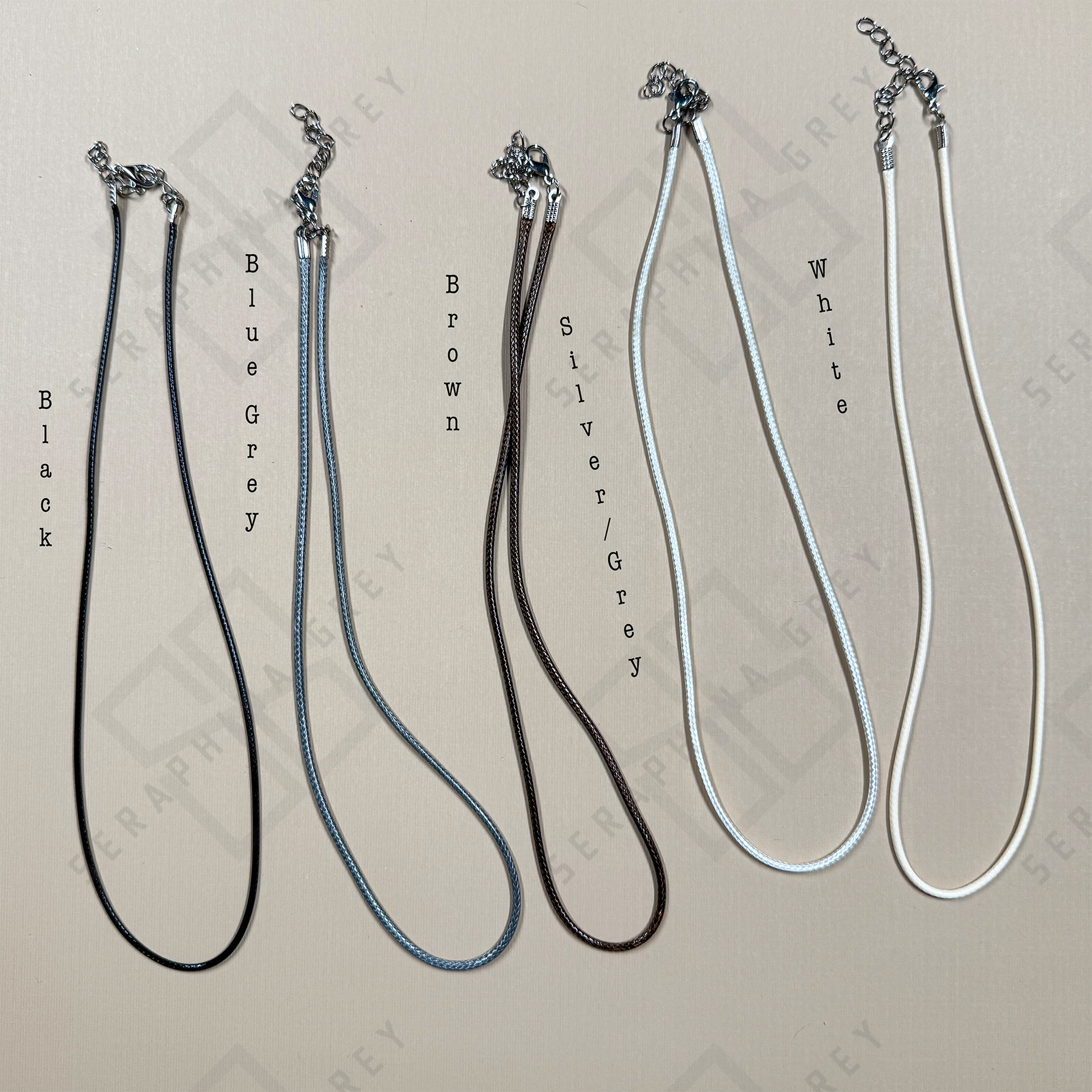 Five adjustable wax cord necklaces displayed side by side in black, blue-grey, brown, silver-grey, and white, each with silver-tone clasps and extender chains.
