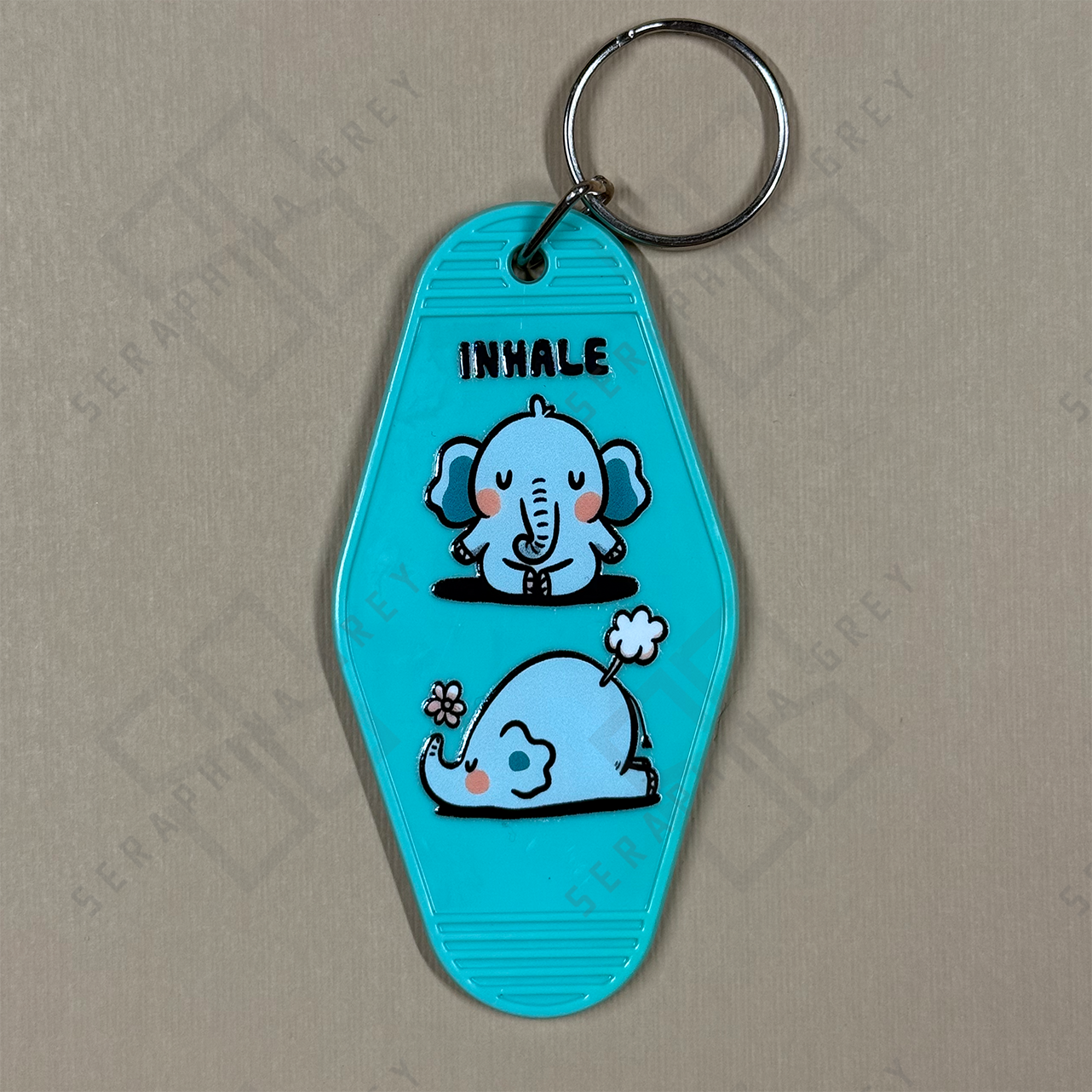 Blue keychain with cartoon elephants and 'INHALE' text , exhale is a fart on a beige background
