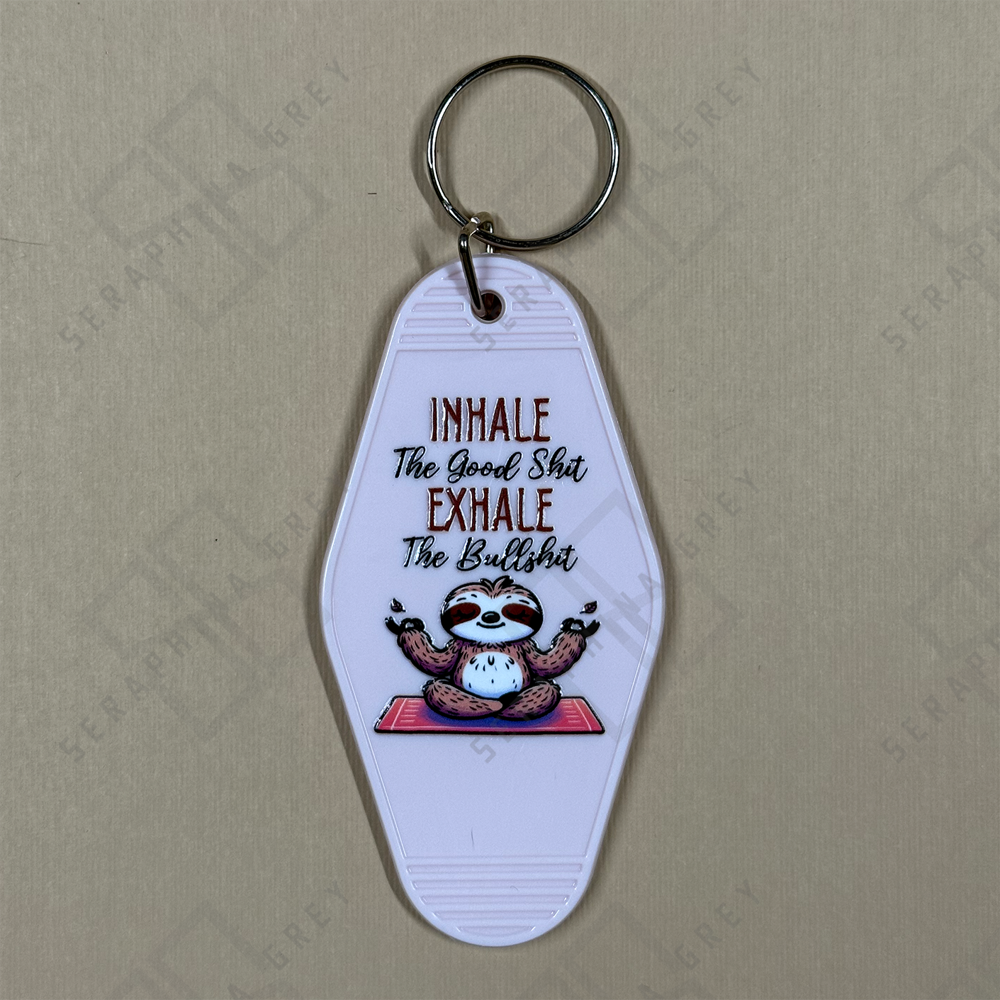 Keychain with sloth design and humorous text Inhale the good sh. exhales the bs. on a beige background