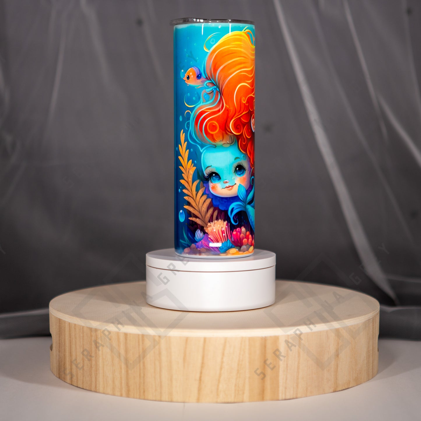 The image shows a side view of a vibrant tumbler featuring a sweet blue face with flowing orange hair peeking out behind her MerMom. They are surrounded by underwater elements such as coral, seaweed, bubbles and small fish near the top. The background is a bright blue, enhancing the underwater theme. The tumbler is placed on a circular wooden stand with a white base, set against a soft-focus gray background. The overall look is whimsical and appealing, ideal for anyone who loves mermaid-themed items.